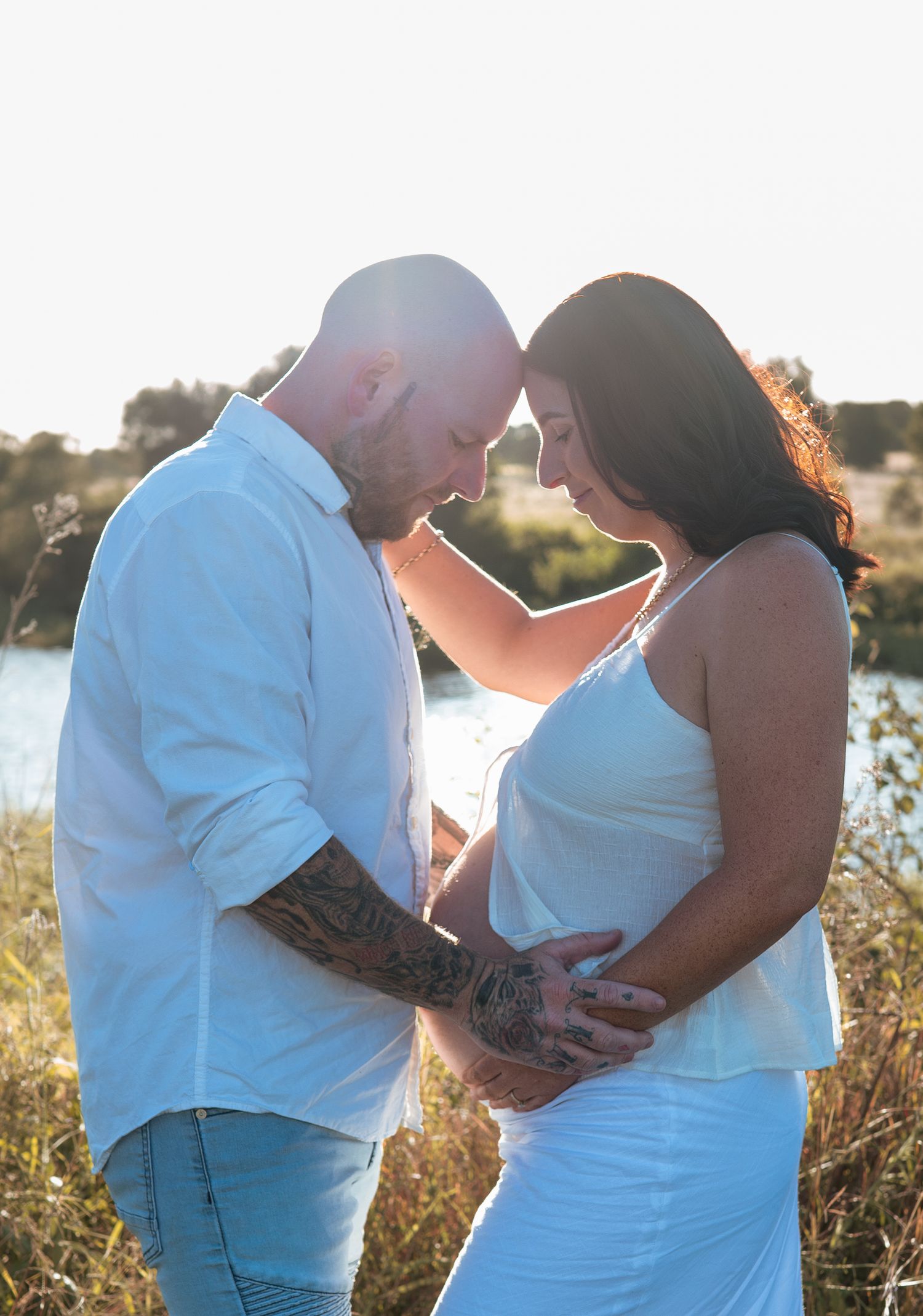 Intimate maternity portrait of couple in white clothing at sunset by the water.