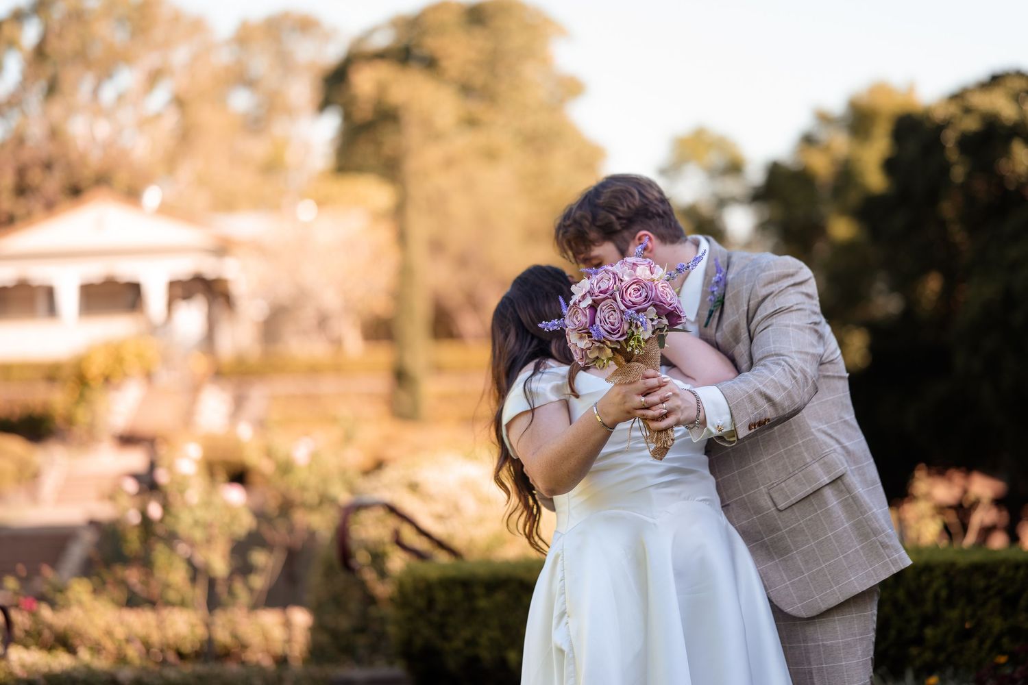 A bride and groom share a kiss while holding a purple bouquet in a sunlit garden wedding setting.