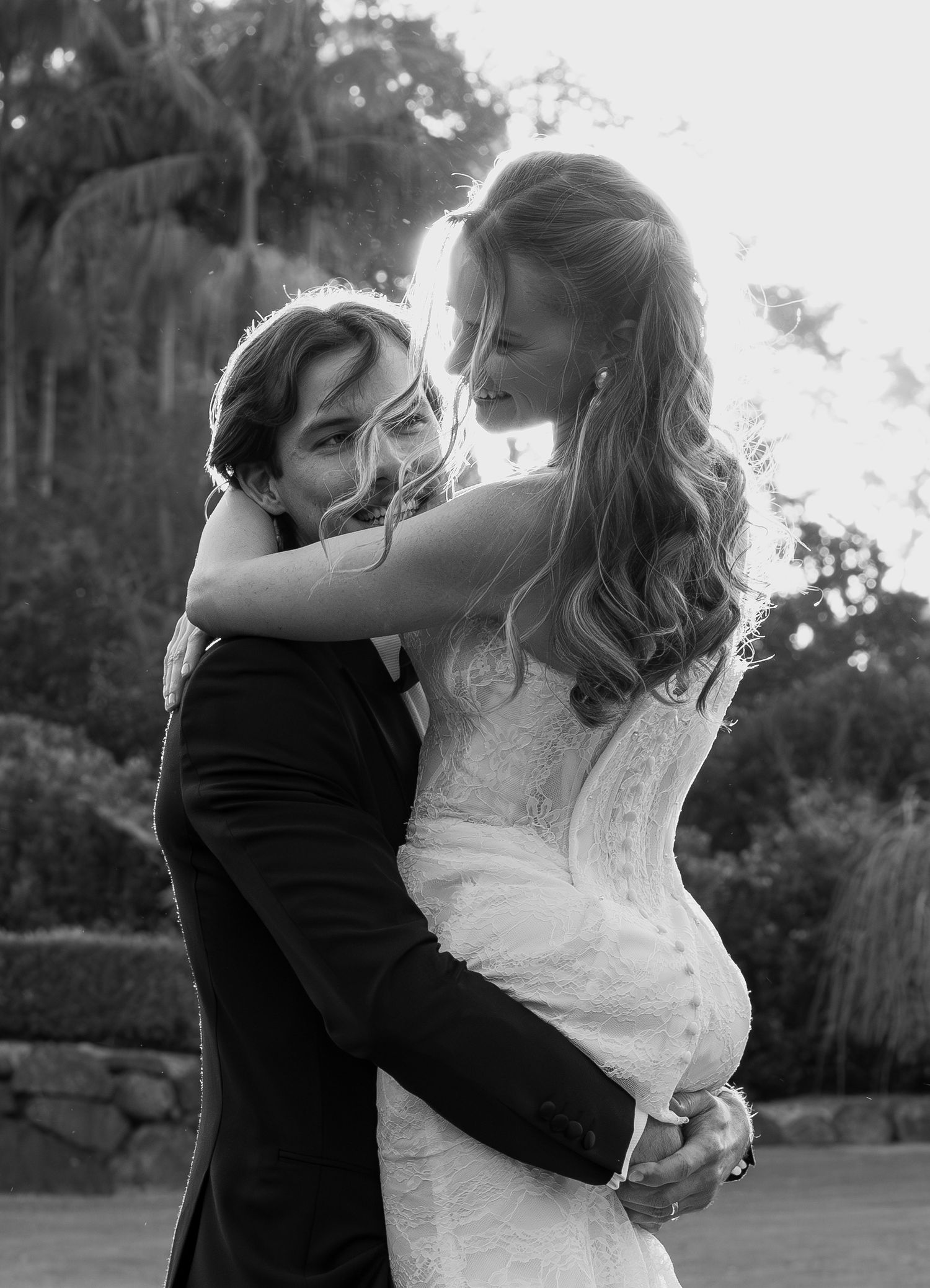 Bride and groom embrace in black and white wedding photo outdoors with trees in background.