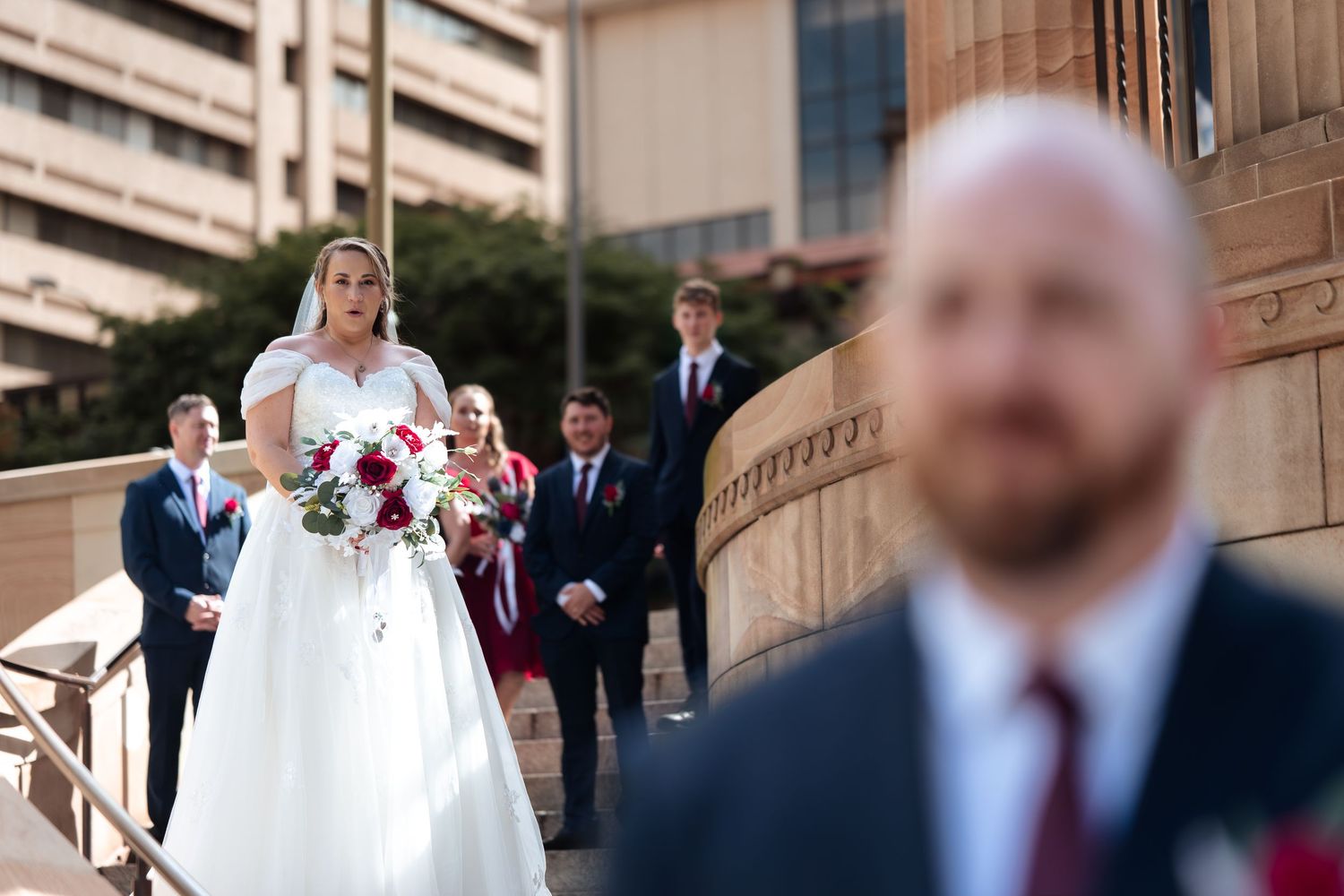 Bride in white gown holds red bouquet on steps as groom stands in foreground with wedding party behind.