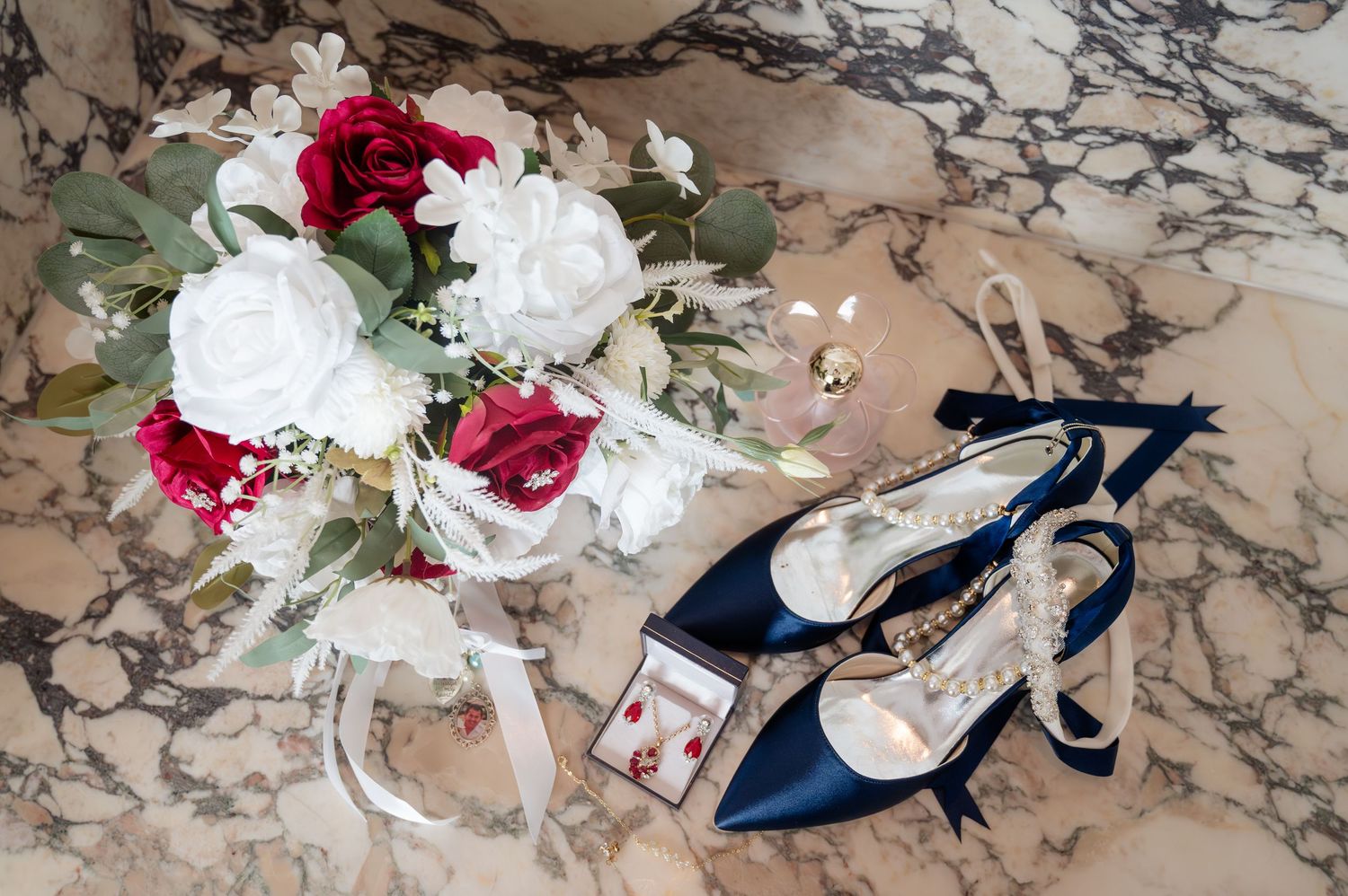 Navy blue bridal heels beside a red and white rose bouquet with greenery and earrings on marble surface.