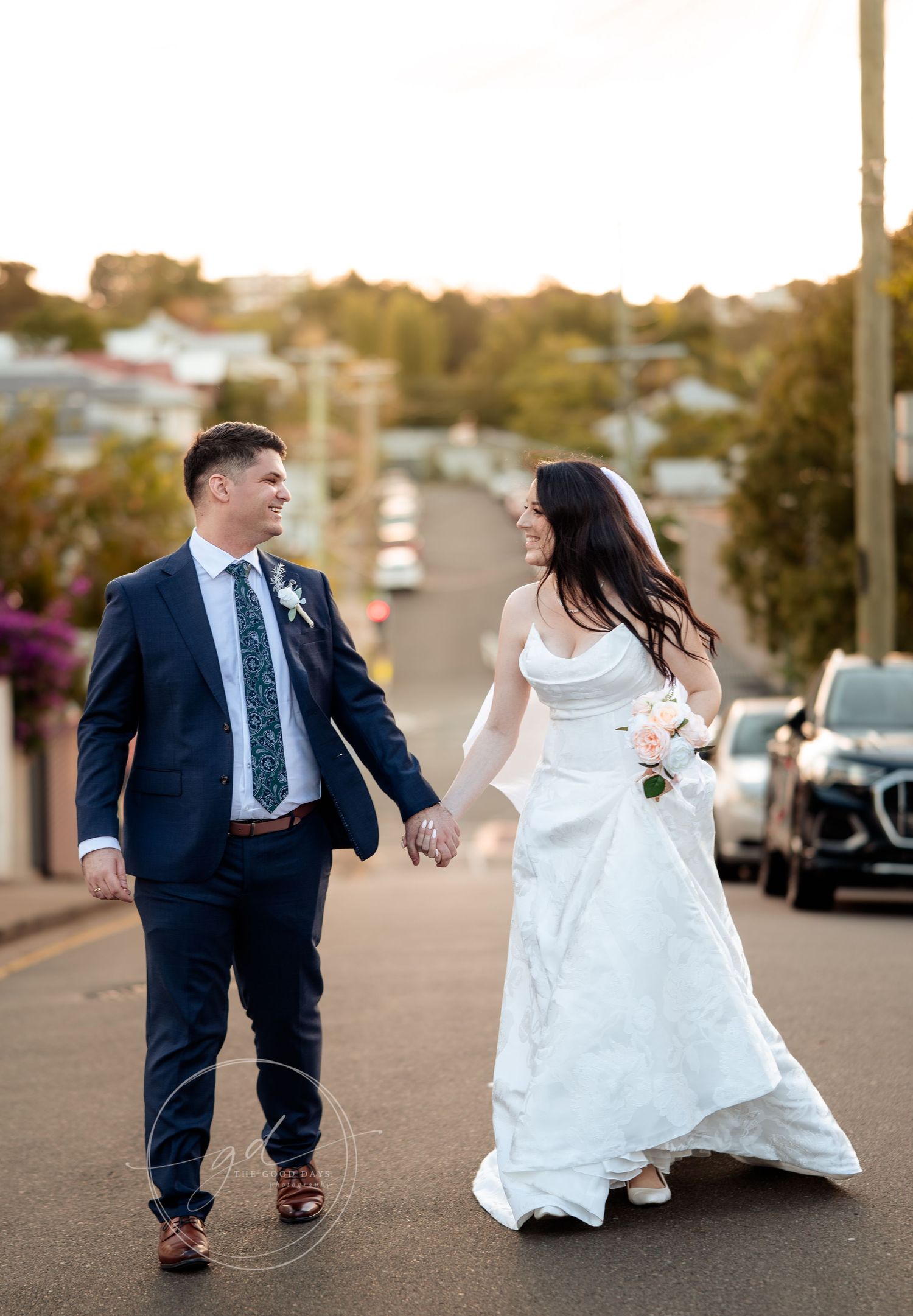 Bride and groom walk hand-in-hand down a sunlit street, she in a white gown, he in a navy suit with floral boutonniere.