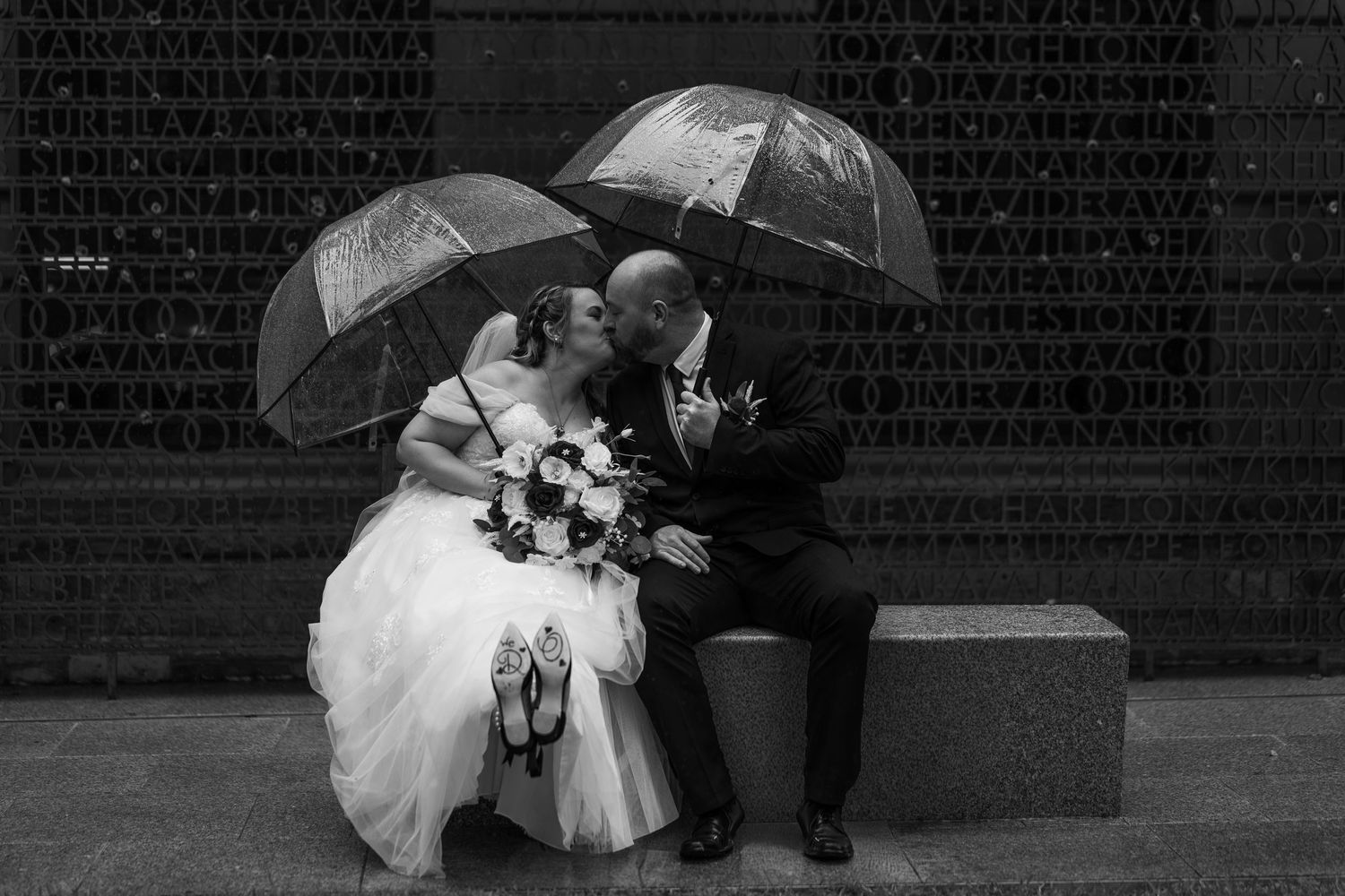 Black and white photo of bride and groom kissing on bench in rain under clear umbrellas.