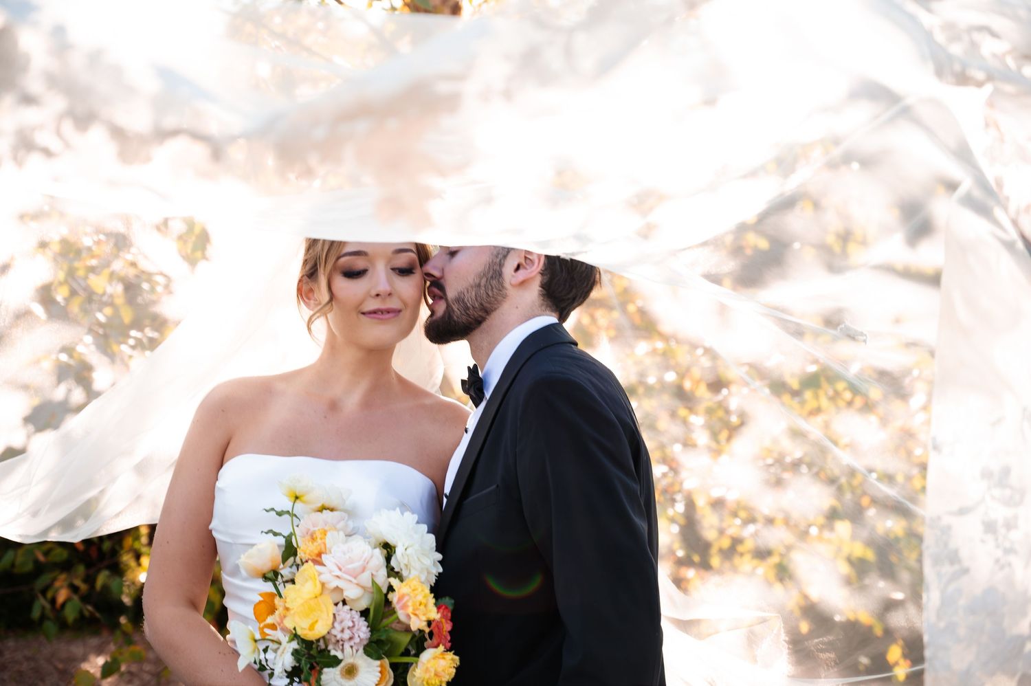 Bride holding yellow bouquet as groom kisses her cheek under flowing veil outdoors.