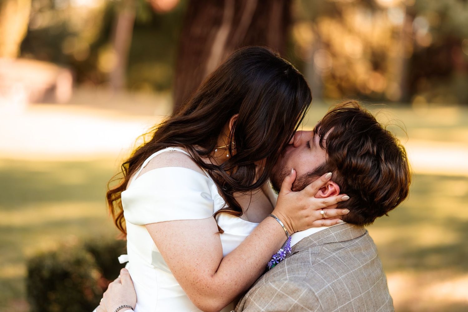 Couple sharing a romantic kiss outdoors in a sunlit park, woman in white dress cradling mans face tenderly.