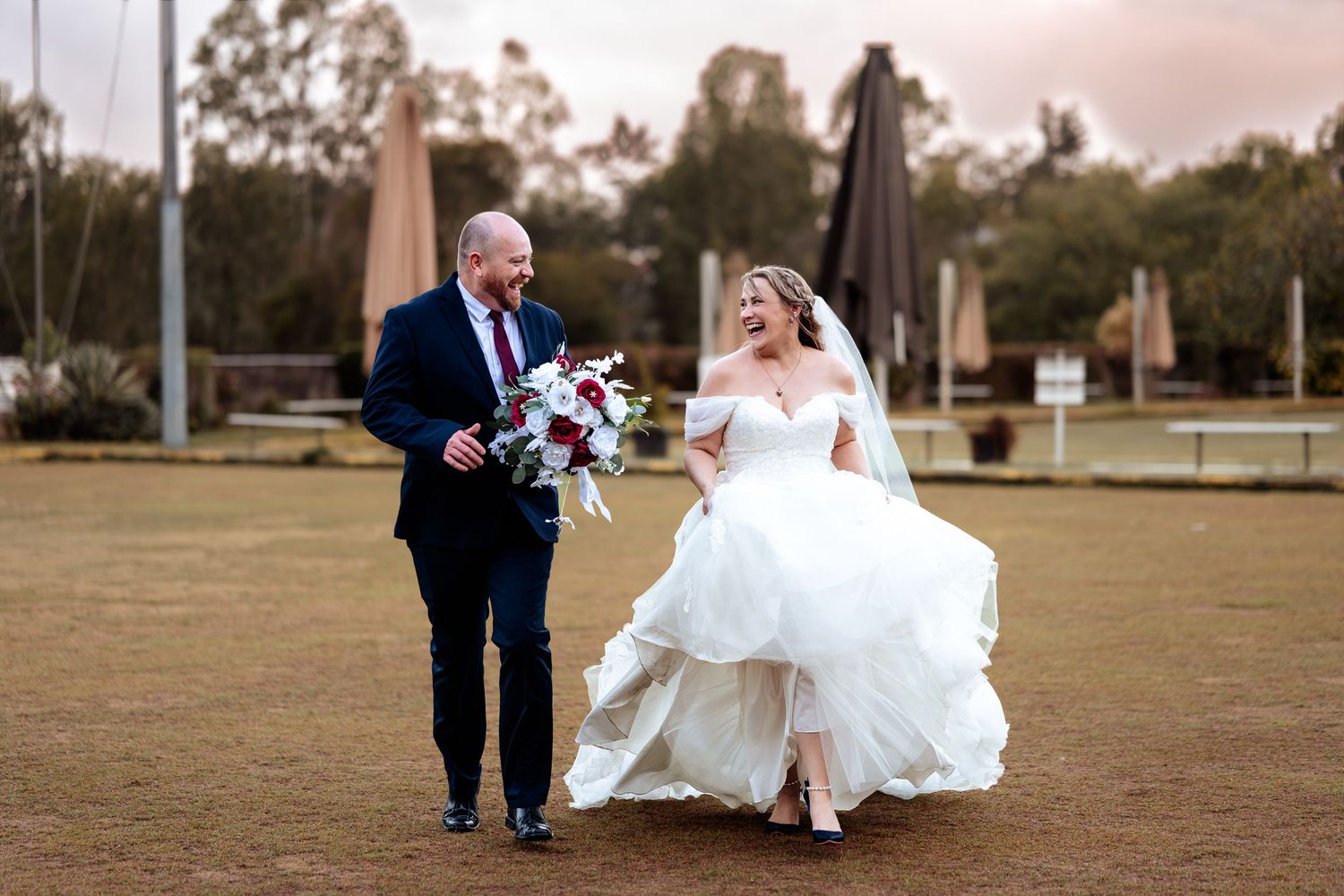 Bride and groom walking outdoors, bride lifting white gown, groom holding bouquet on wedding day.