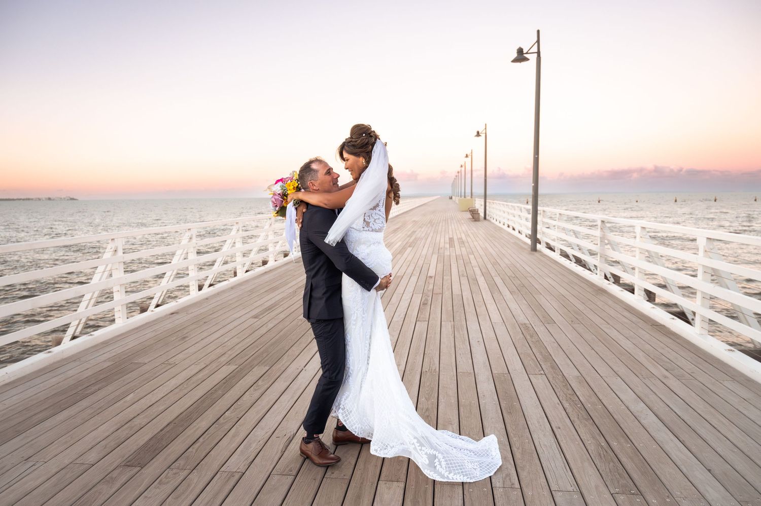 Couple sharing romantic embrace on wooden pier at sunset, bride in white gown lifted by groom in navy suit.