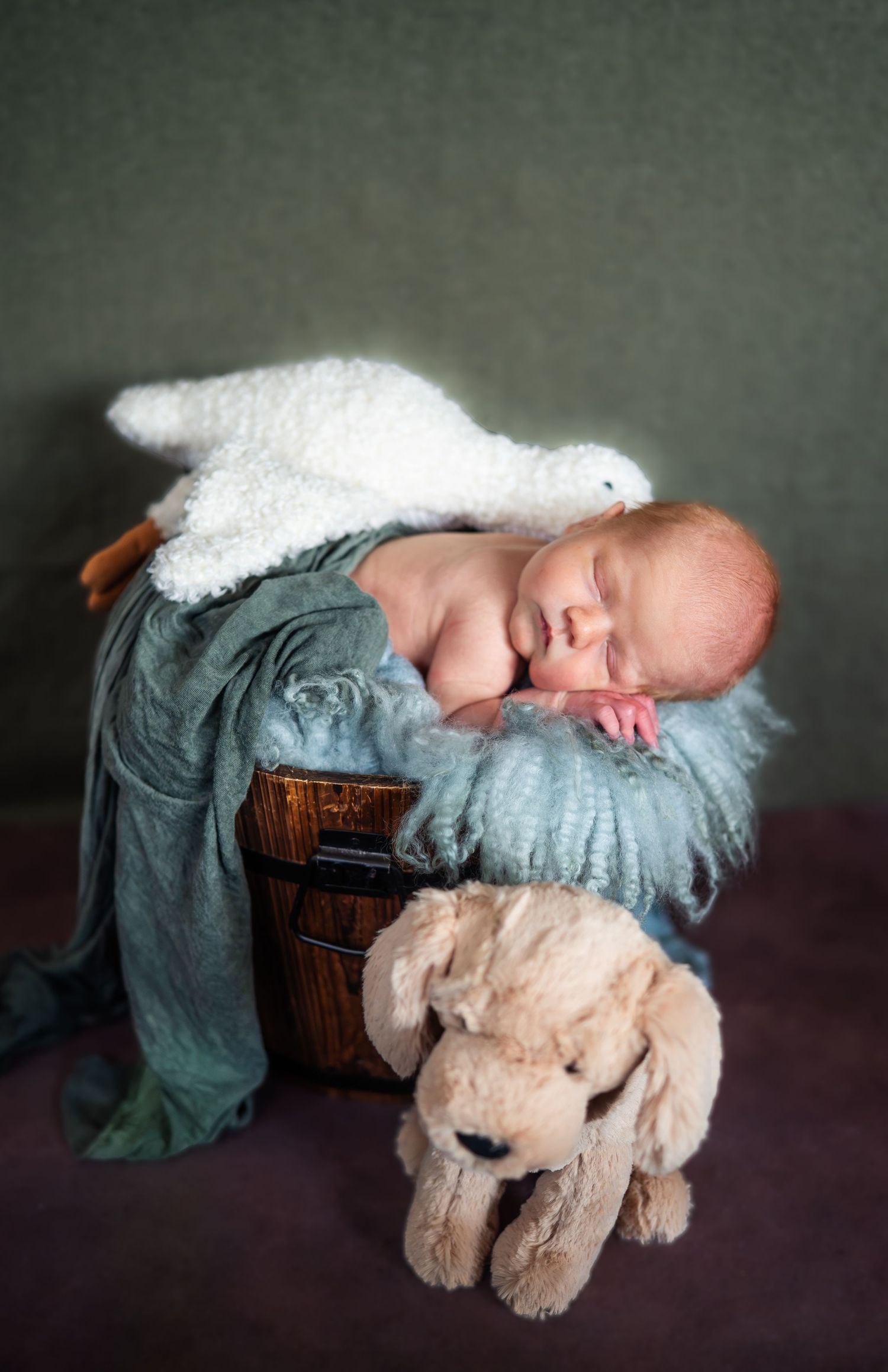 Sleeping newborn in blue blanket rests peacefully next to plush teddy bear on wooden surface.