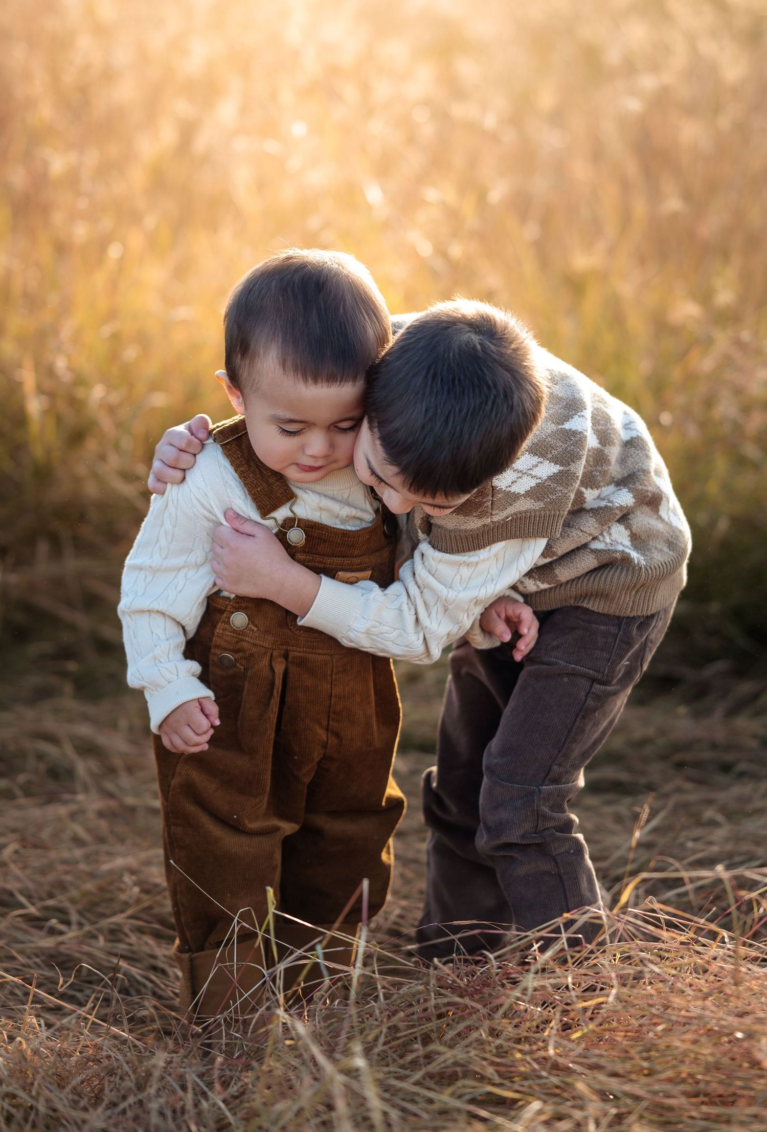 Two young children in brown overalls sharing a sweet moment in autumn field.