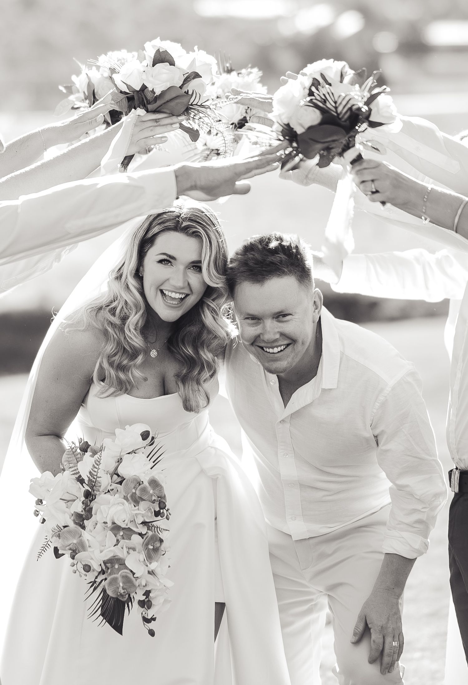 Black and white photo of a smiling bride and groom walking under an arch of bouquets held by wedding party members.