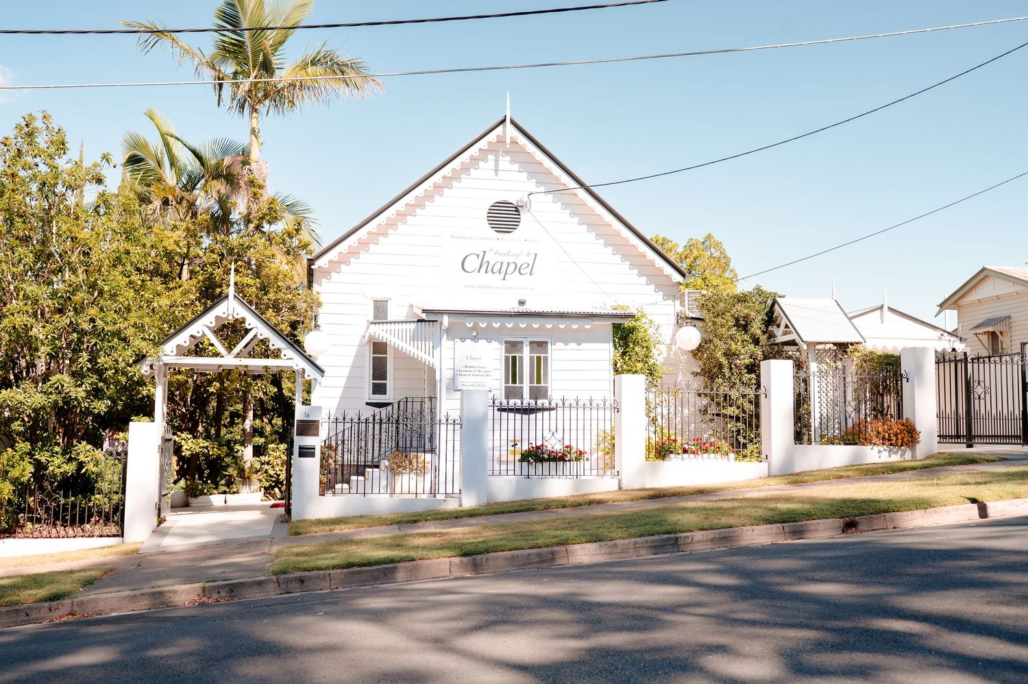 White chapel wedding venue with decorative gate surrounded by lush trees on a sunny day.
