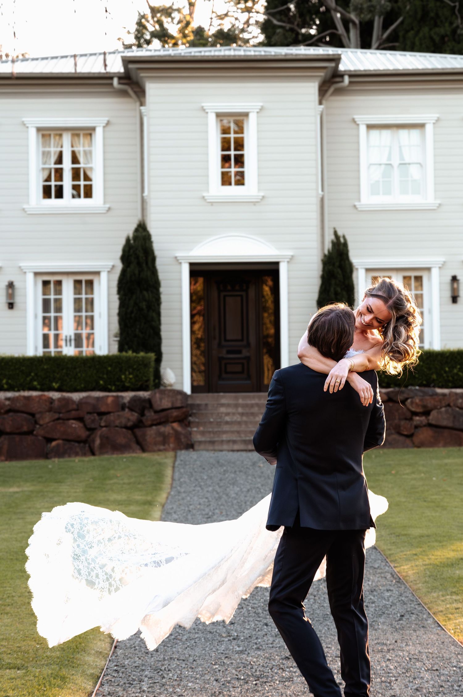 Groom carries bride in flowing white wedding dress in front of elegant white house with dark wooden door.