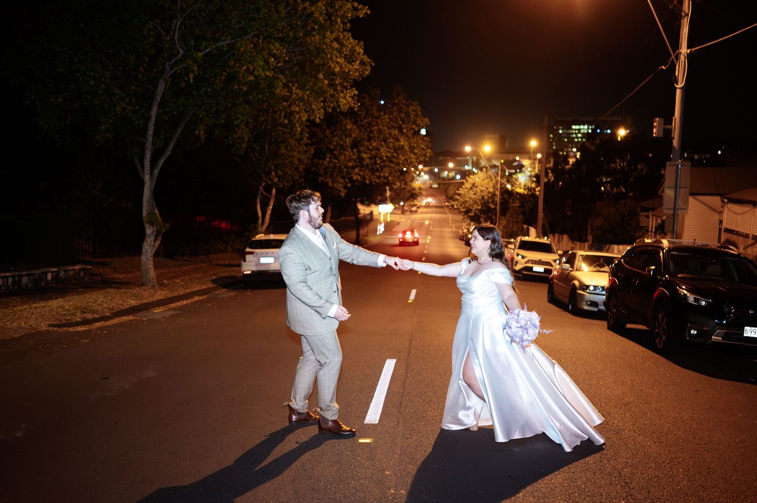 Bride and groom dancing on a street at night, with city lights and parked cars in the background.