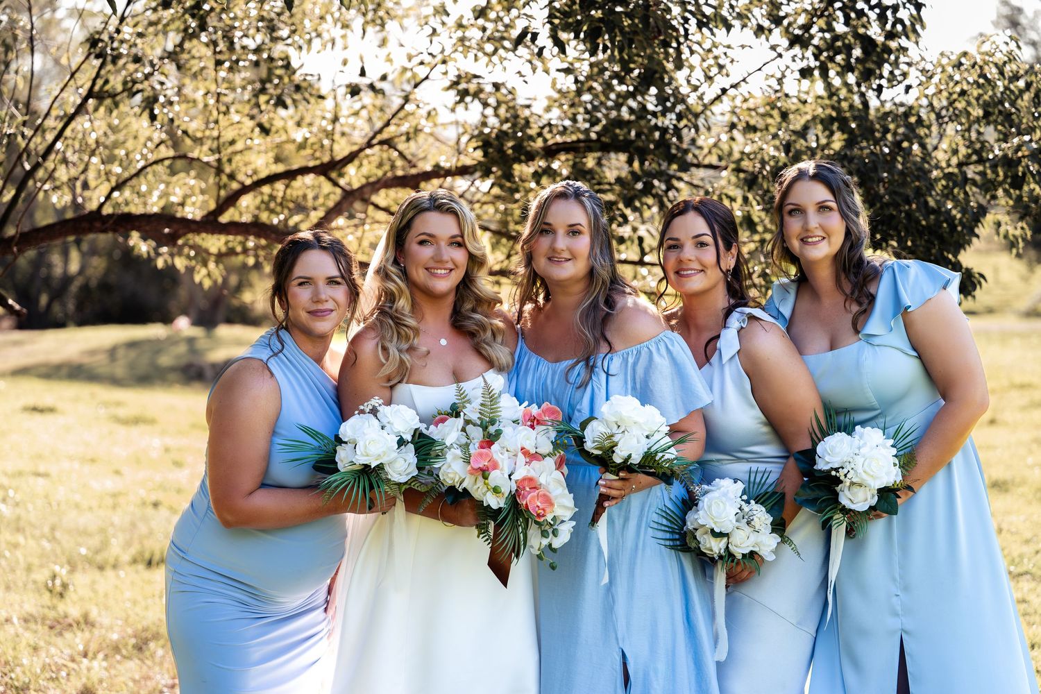 Bride in white gown with four bridesmaids in light blue dresses holding white and pink bouquets outdoors.