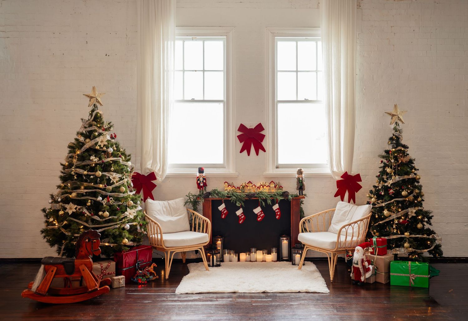 Festive living room with two decorated Christmas trees, rattan chairs, fireplace with stockings, and red bows.