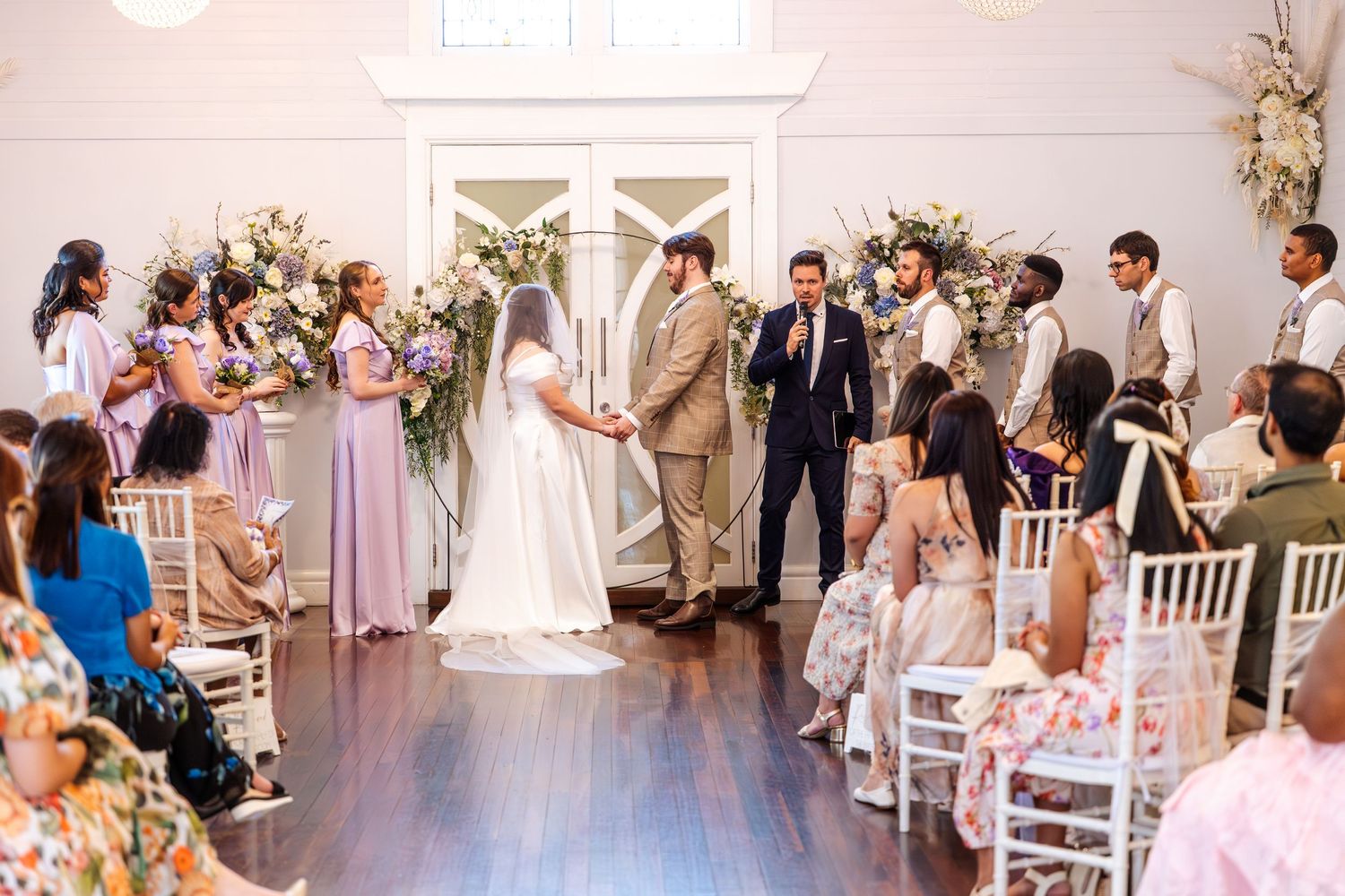 Elegant indoor wedding ceremony with bride and groom at altar, bridesmaids in lavender, floral arch, white chairs, wood floors.