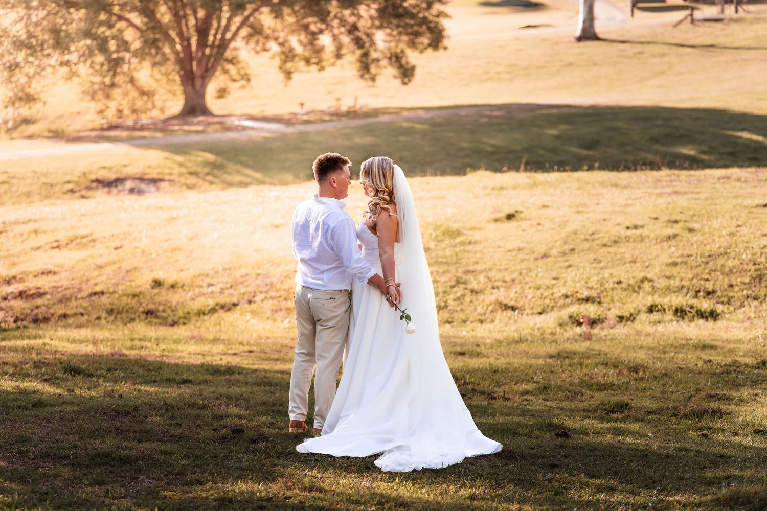 Bride and groom stand together on a sunlit green lawn with warm golden hour light behind them.