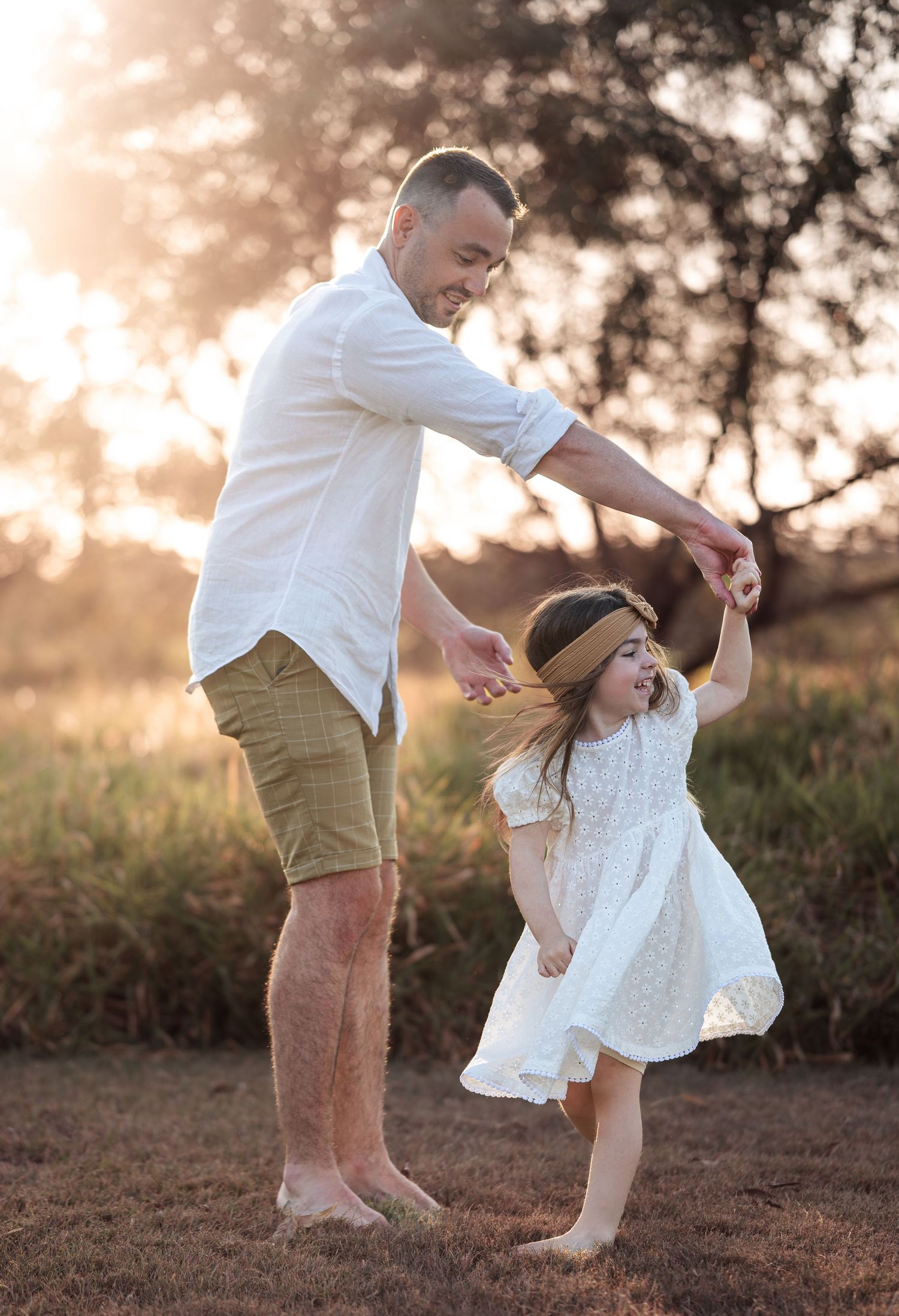 Parent and child dancing together in a field at golden hour wearing white and khaki outfits.