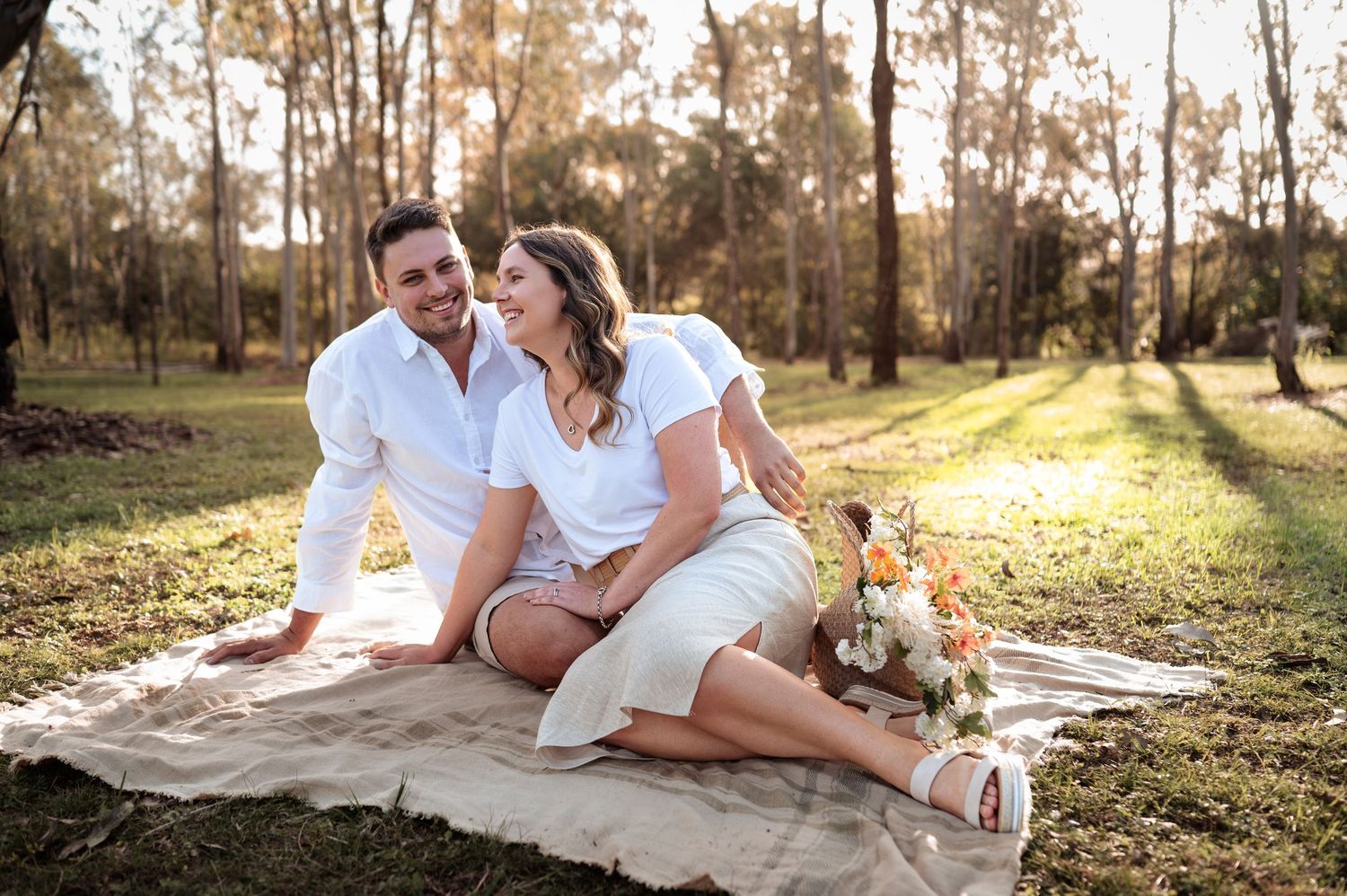A romantic outdoor photoshoot with a couple laying on a blanket in a wooded setting at golden hour.