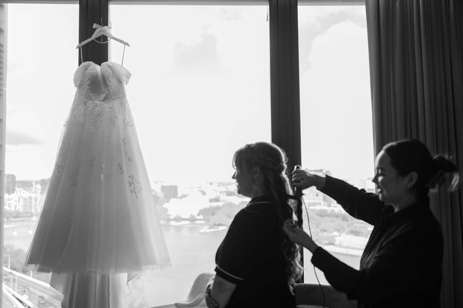 Black and white photo of bride getting hair done with wedding dress hanging by window.