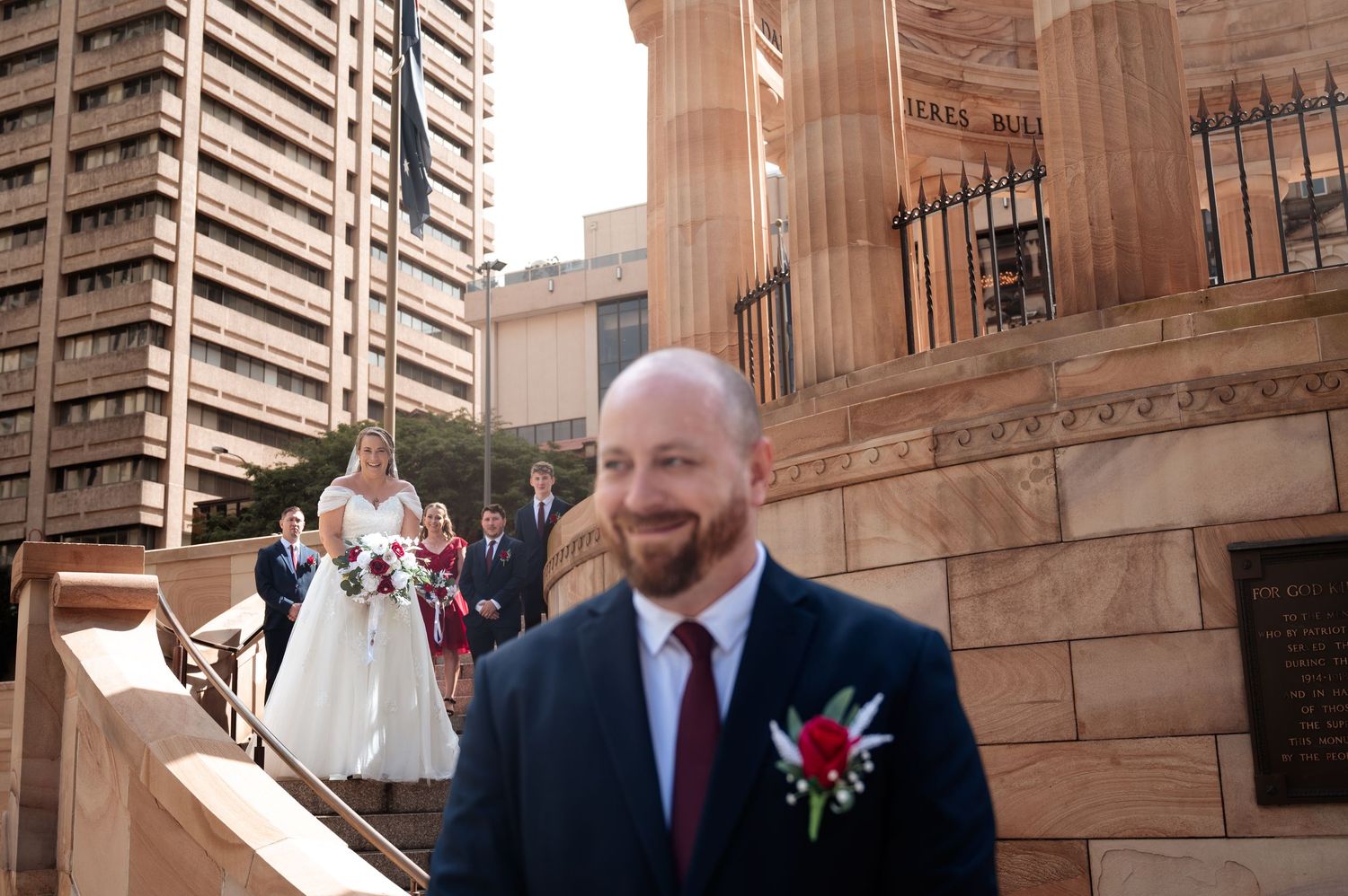 Groom smiling in foreground with wedding party descending stone church steps in urban setting.
