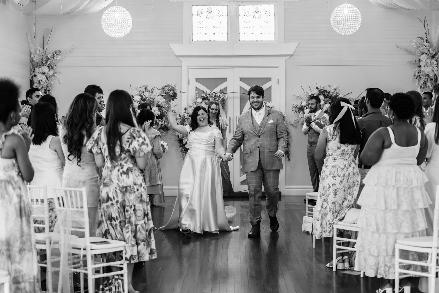 Newlywed couple walks back down aisle as guests cheer in elegant white wedding venue ceremony.