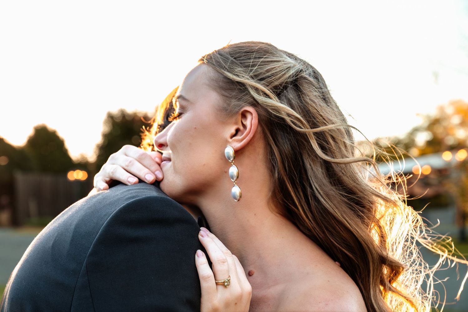 Bride with pearl drop earrings embracing groom at golden hour outdoor wedding ceremony.