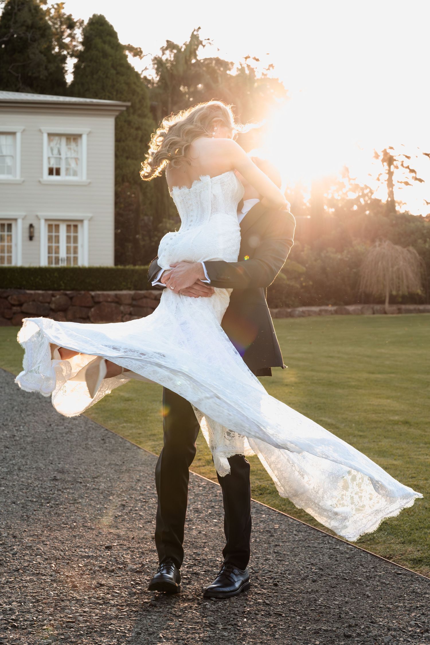 Groom lifting bride in white wedding gown at golden hour, elegant estate in background.
