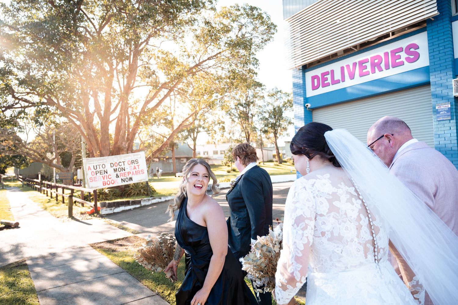Bride in lace gown laughing with guests outside a deliveries building on a sunny wedding day.