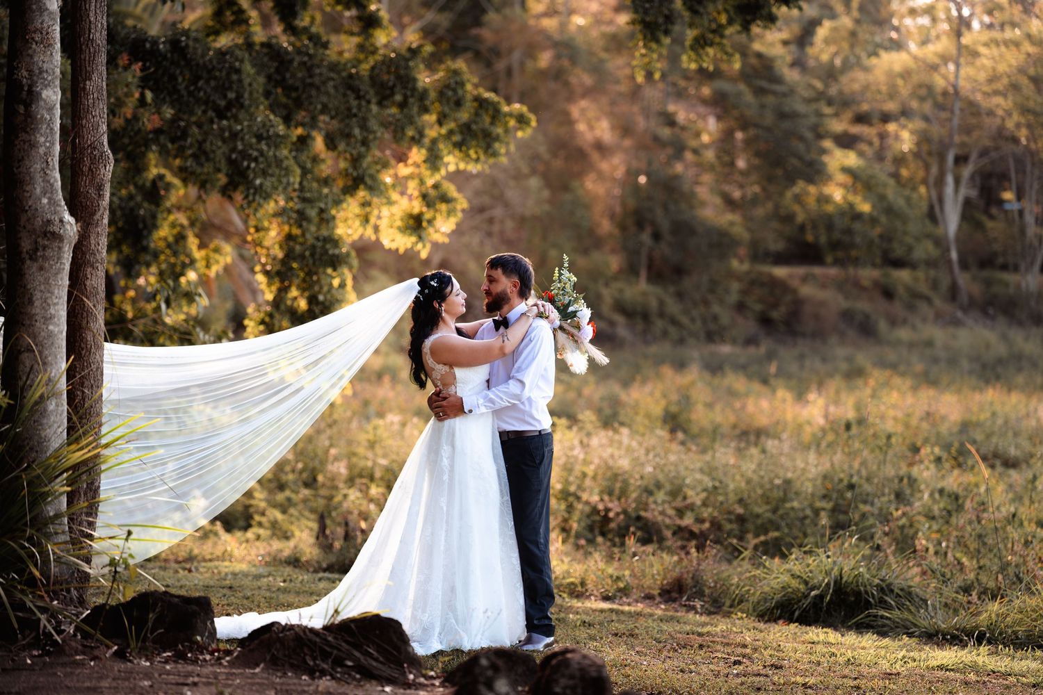 Bride and groom share a romantic kiss outdoors, with a flowing veil caught in the breeze at a garden wedding.