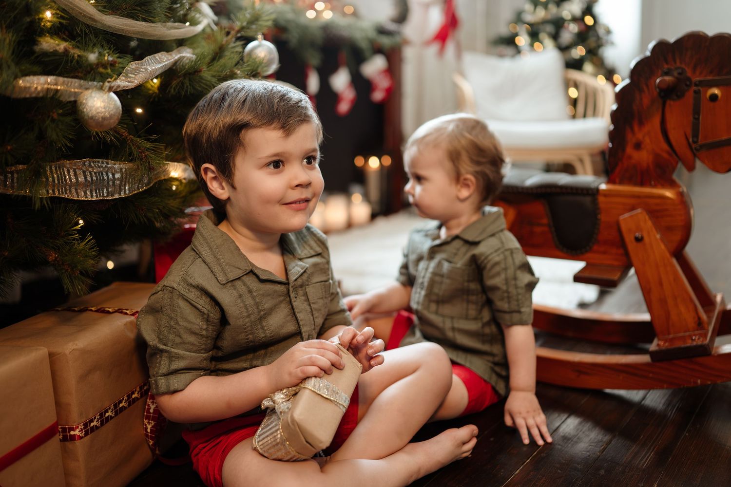 Two young boys in matching outfits opening gifts beside a decorated Christmas tree.