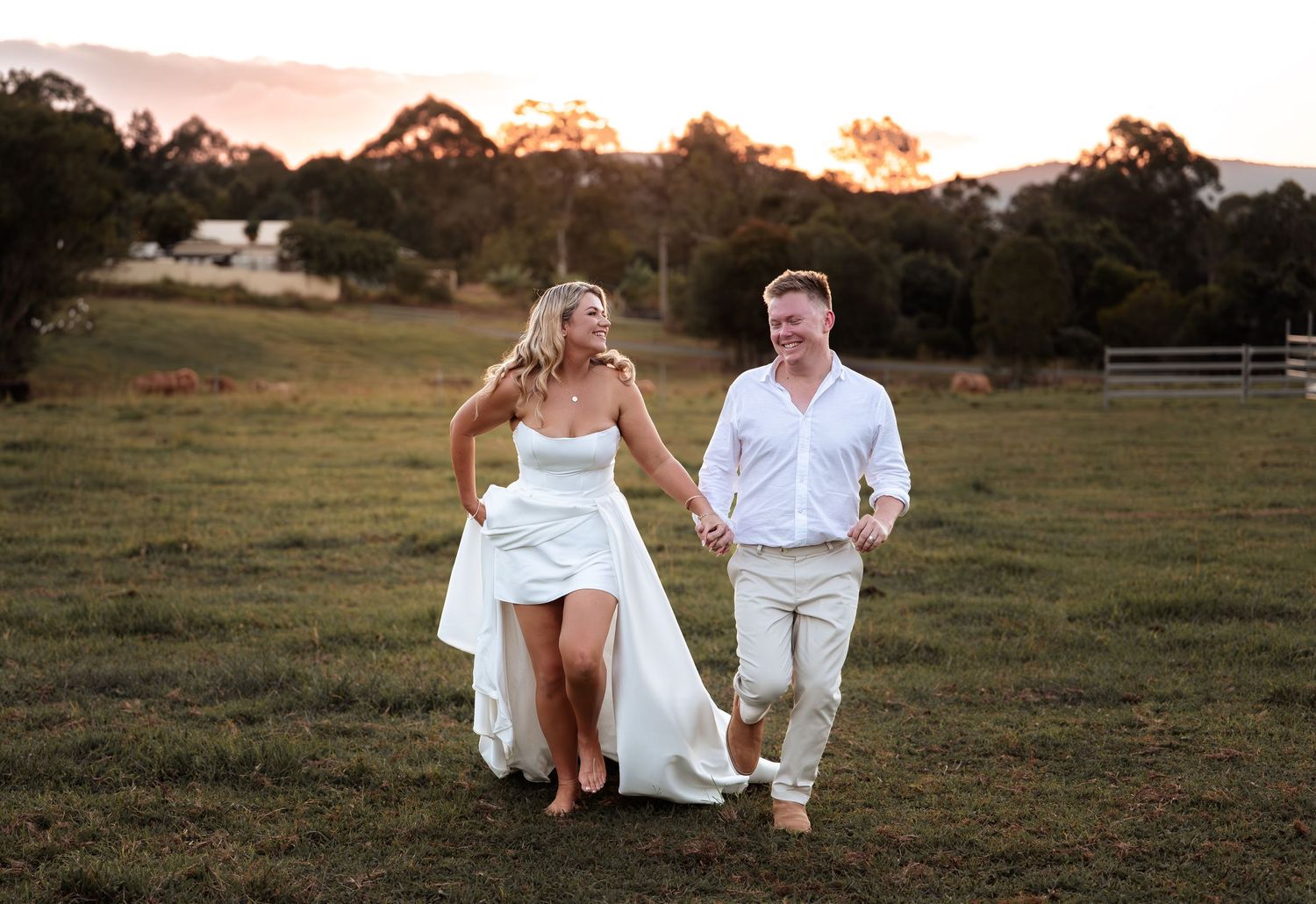 Bride and groom holding hands walking through green field at sunset in white wedding attire.