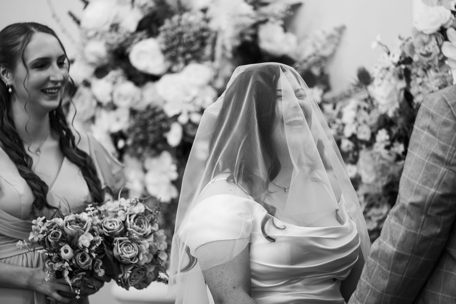 Black and white wedding photo of a veiled bride smiling beside a bridesmaid holding a bouquet near floral arrangements.