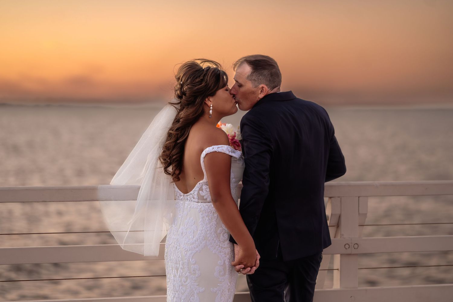 Bride and groom share a romantic kiss on a pier at sunset, with the ocean glowing orange behind them.