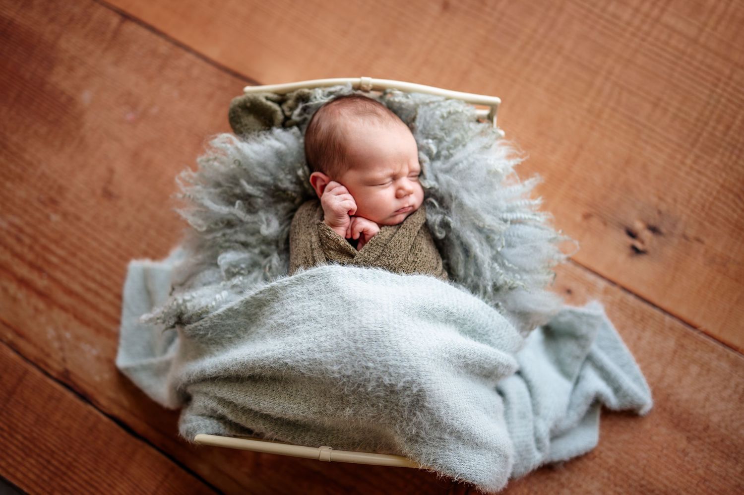 Newborn sleeping on wooden floor wrapped in soft gray blanket.