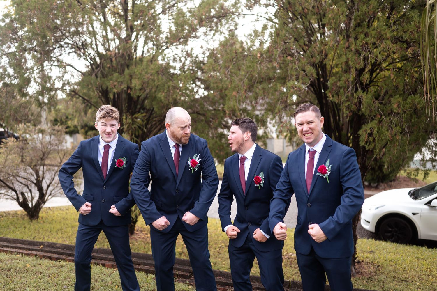 Four groomsmen in navy suits with red boutonnieres strike fun poses outdoors at a wedding.