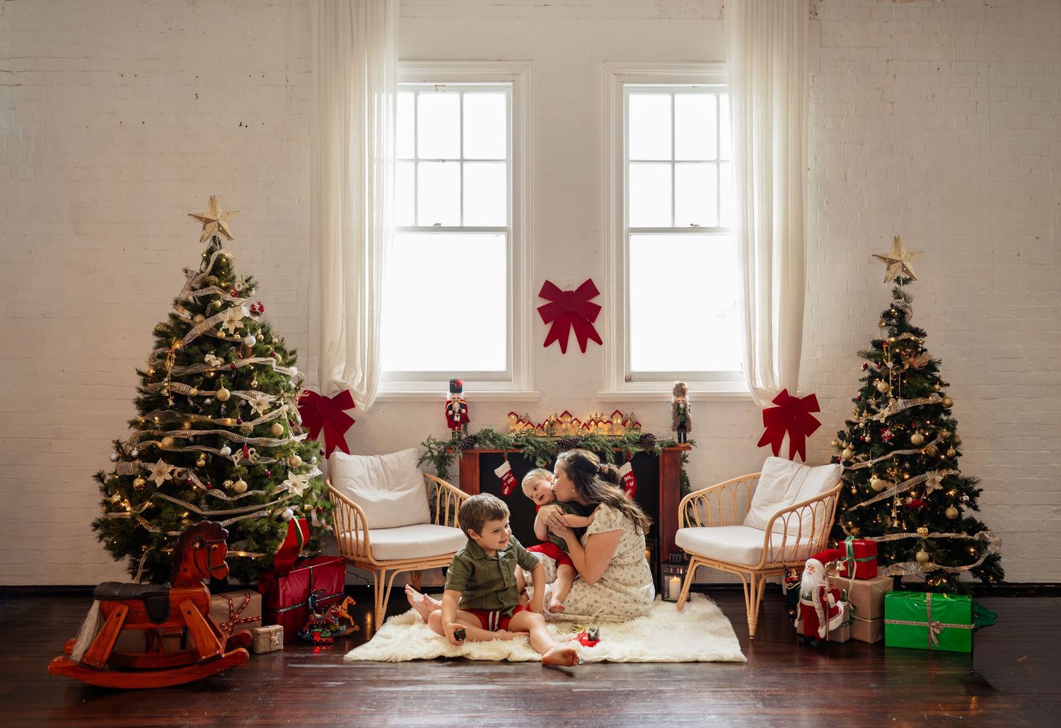 Family celebrating Christmas indoors with decorated trees, gifts, and festive red bows near a fireplace.