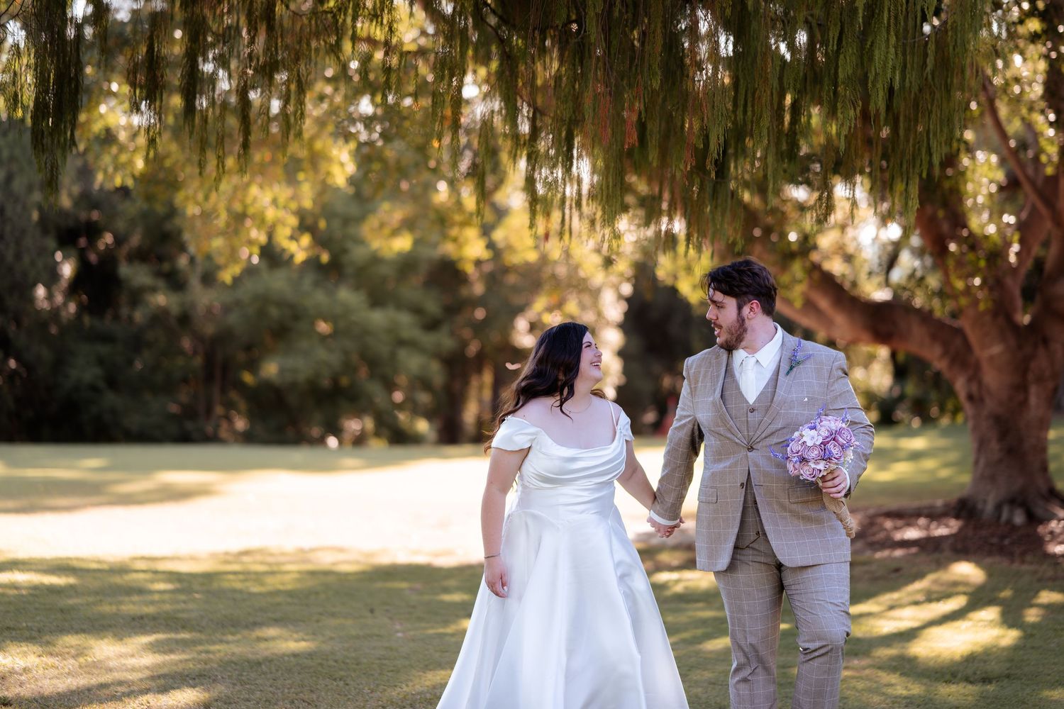 Bride and groom holding hands under weeping willow tree in lush green park, groom holding pink bouquet.