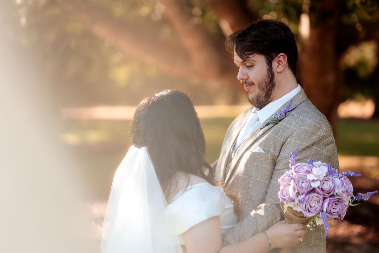 Groom in a beige suit gazes lovingly at bride holding a purple rose bouquet during an outdoor wedding portrait.