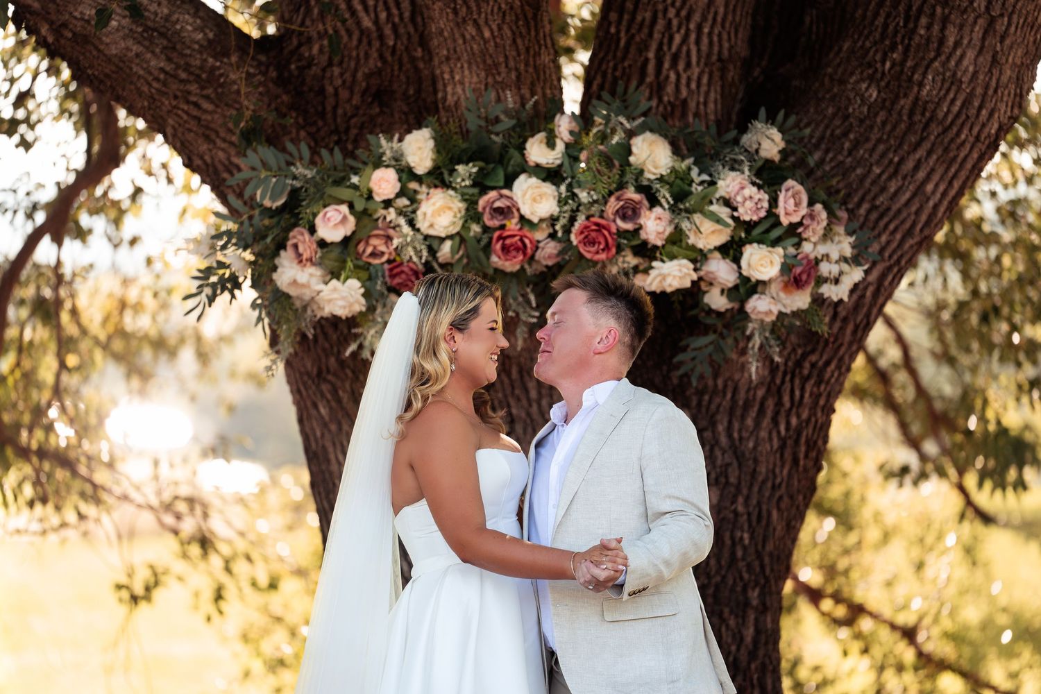 Bride and groom share a romantic kiss under a flower-adorned oak tree during outdoor wedding ceremony.