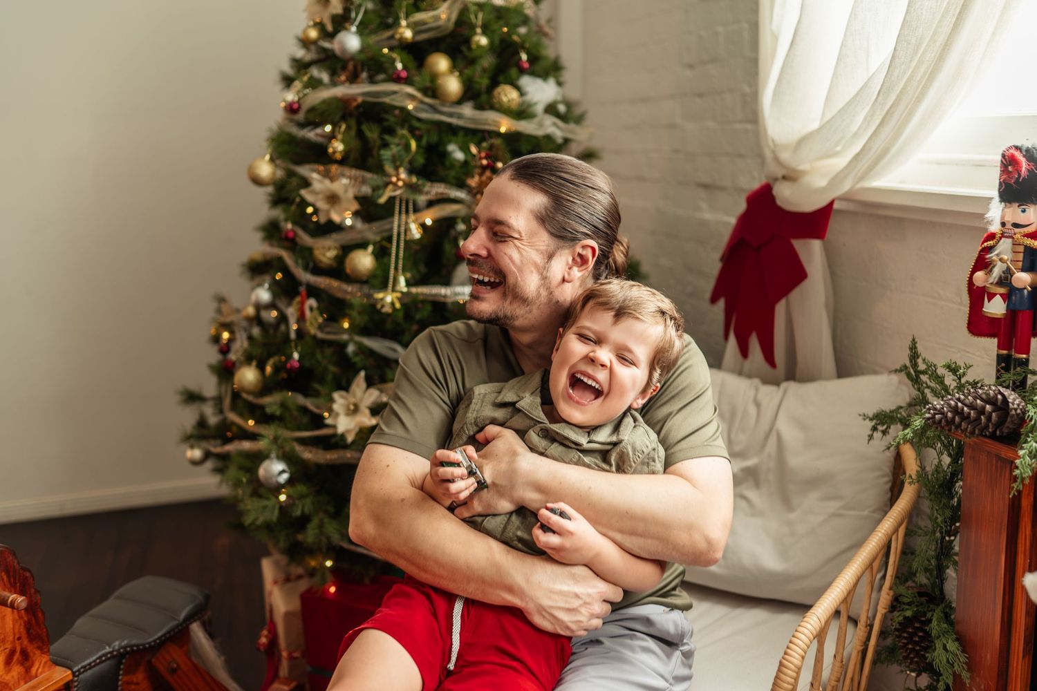 Father laughing and hugging his young son near a decorated Christmas tree in a festive living room.