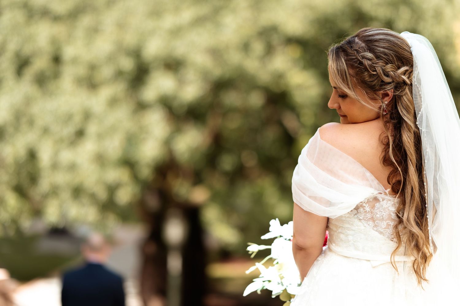Bride in elegant white gown with veil and floral bouquet, hair in loose curls, standing outdoors at wedding ceremony.