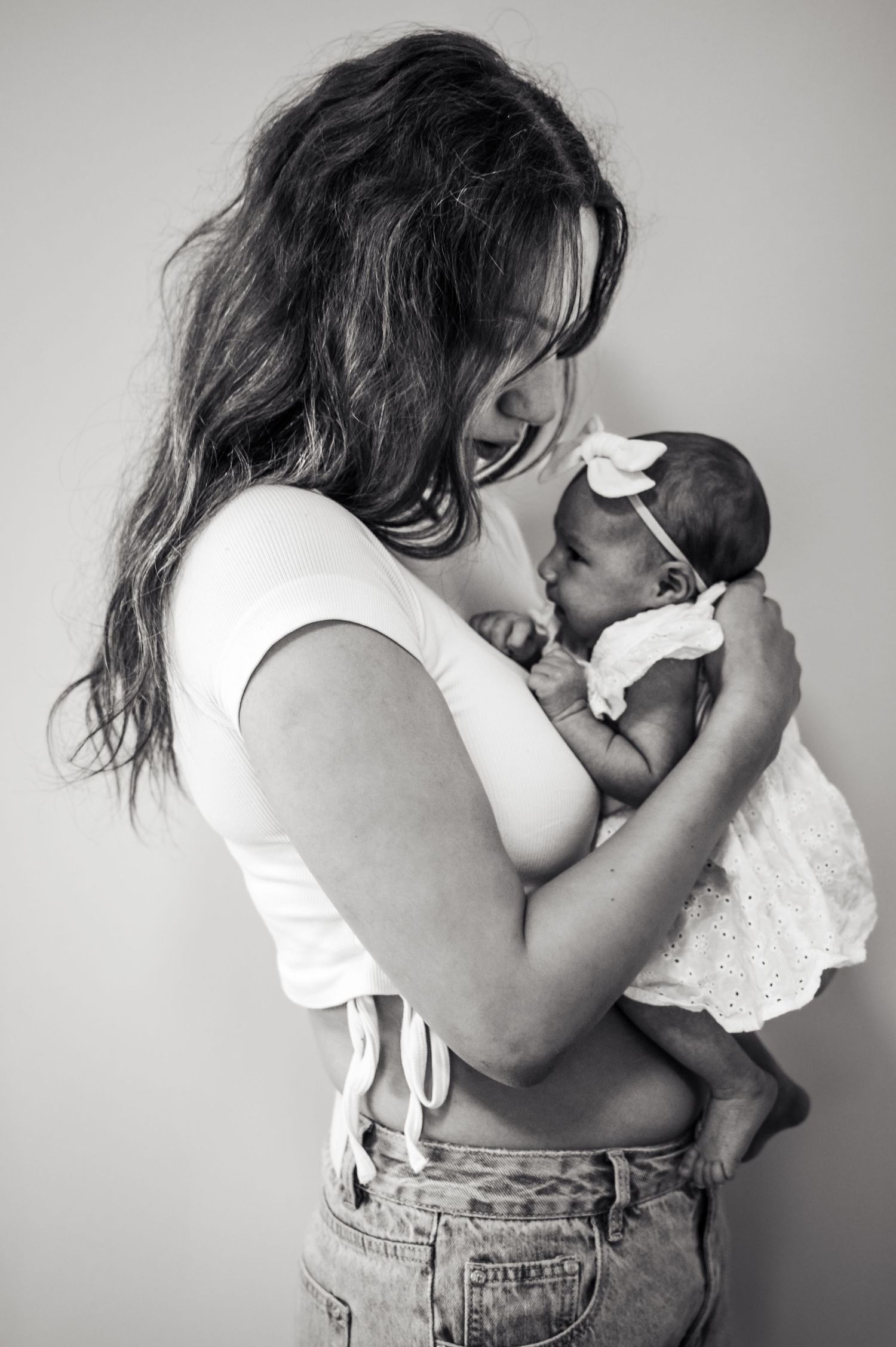 Mother in white top and jeans lovingly embraces newborn baby in black and white photo series.