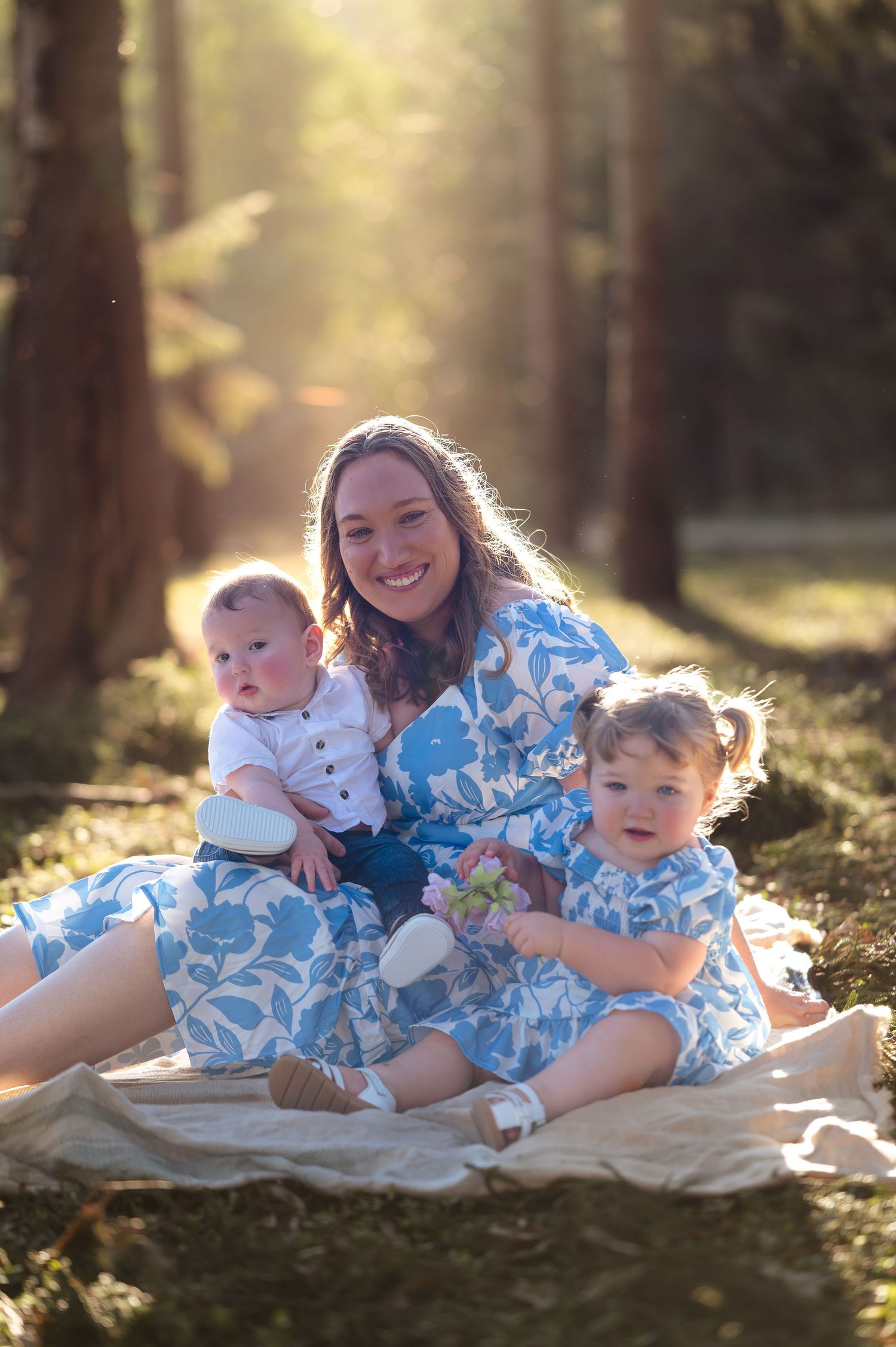 Family group wearing coordinated blue floral outfits posing on blanket in sunlit forest.