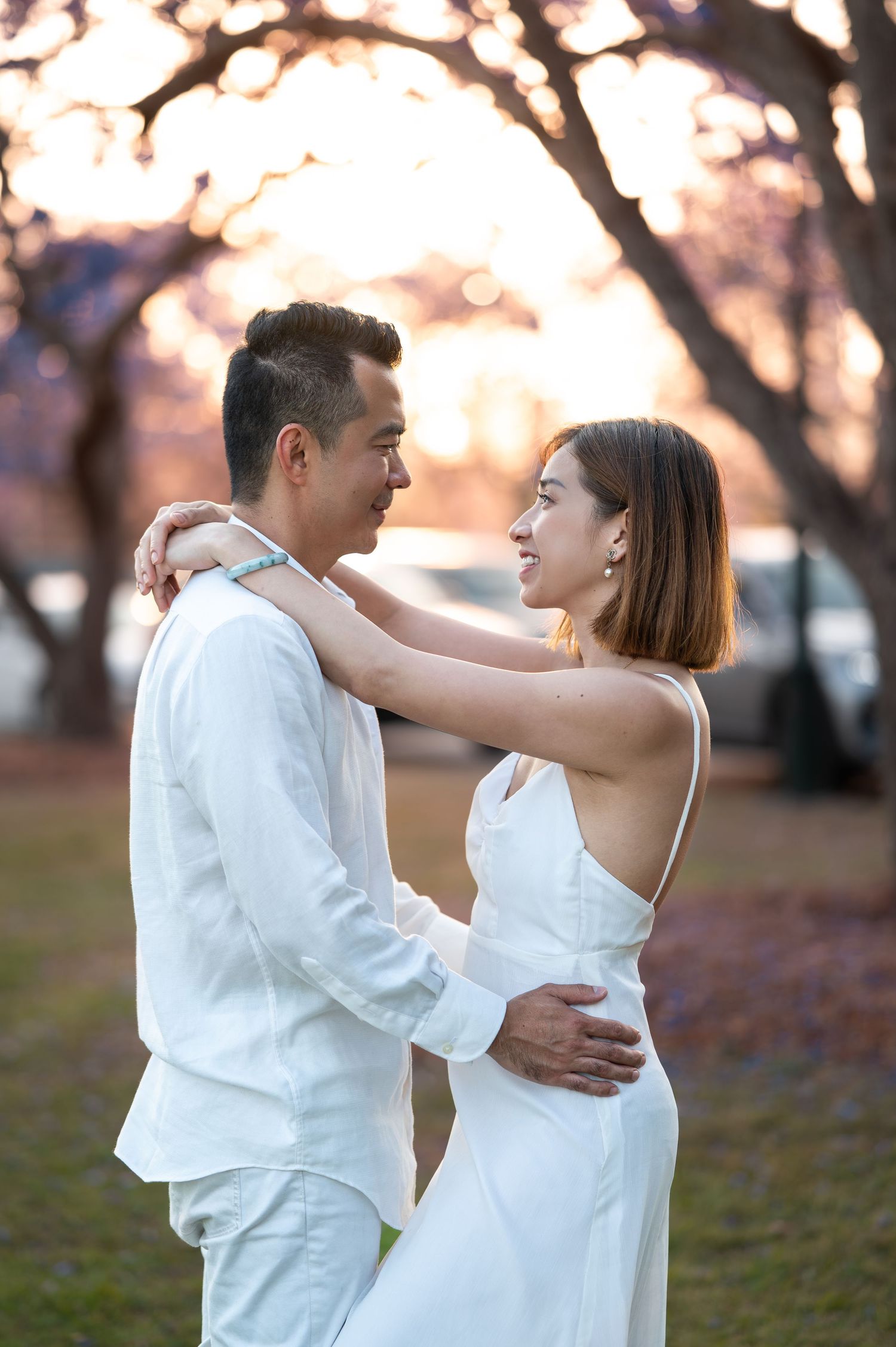 Couple in white formal wear embrace during sunset under cherry blossom trees.