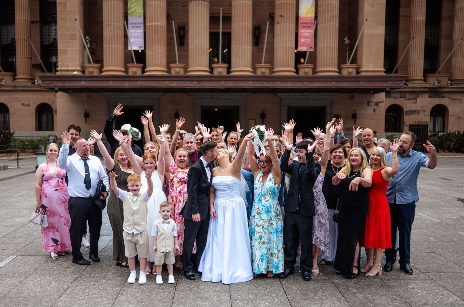 Large wedding party poses together outdoors in front of a brick building with formal attire and colorful dresses.