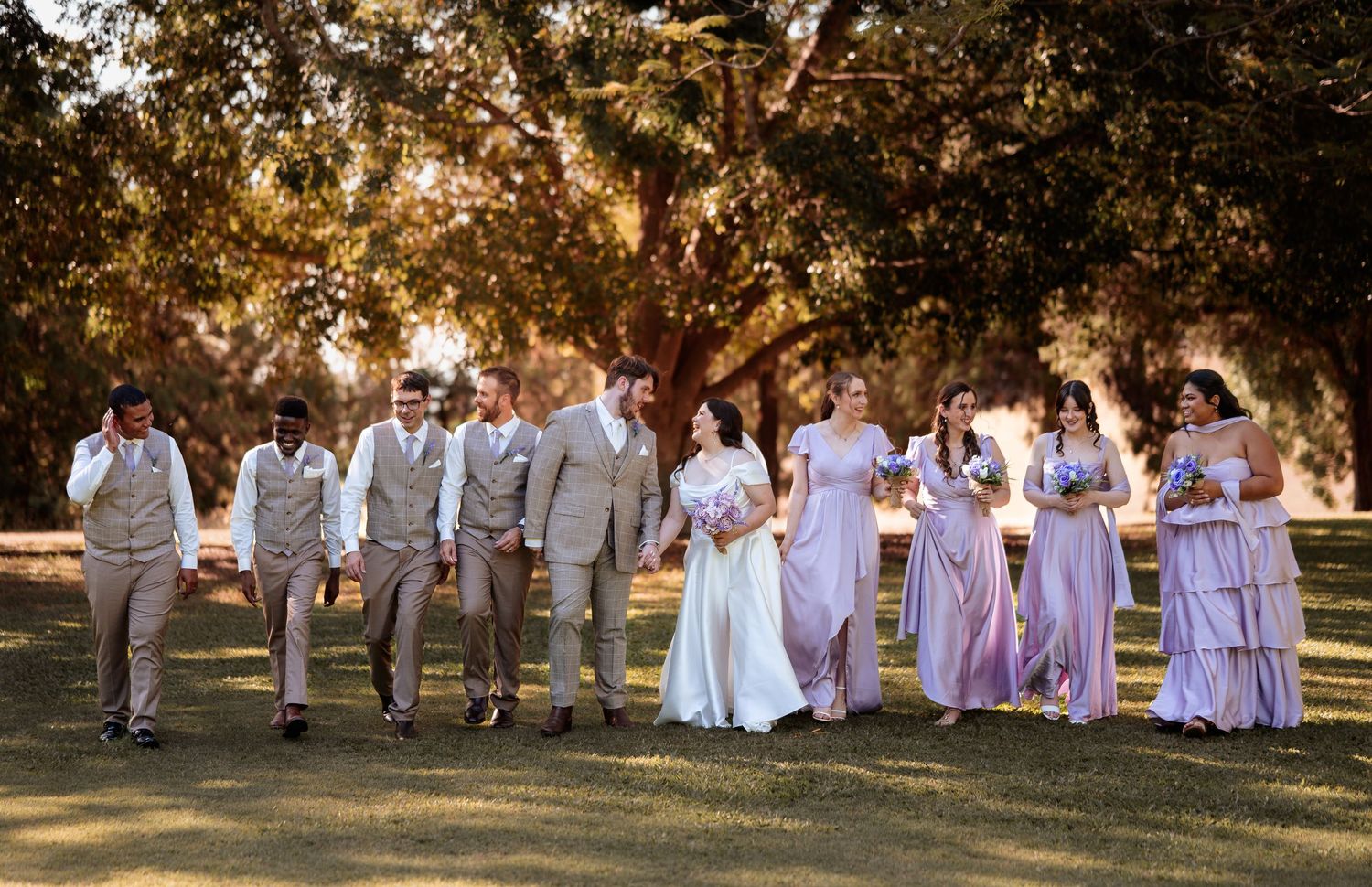 Wedding party walks outdoors in sunlight, groomsmen in tan suits, bridesmaids in lavender gowns, bride in white dress.