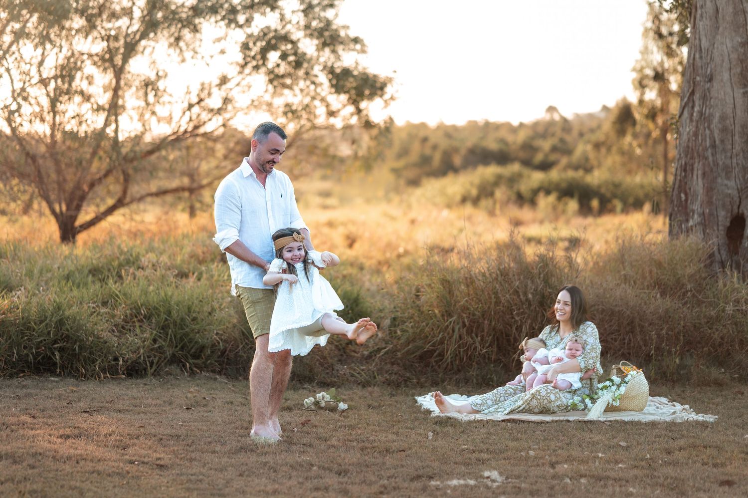 Family photoshoot setup in nature with picnic blanket and rustic decor at sunset.