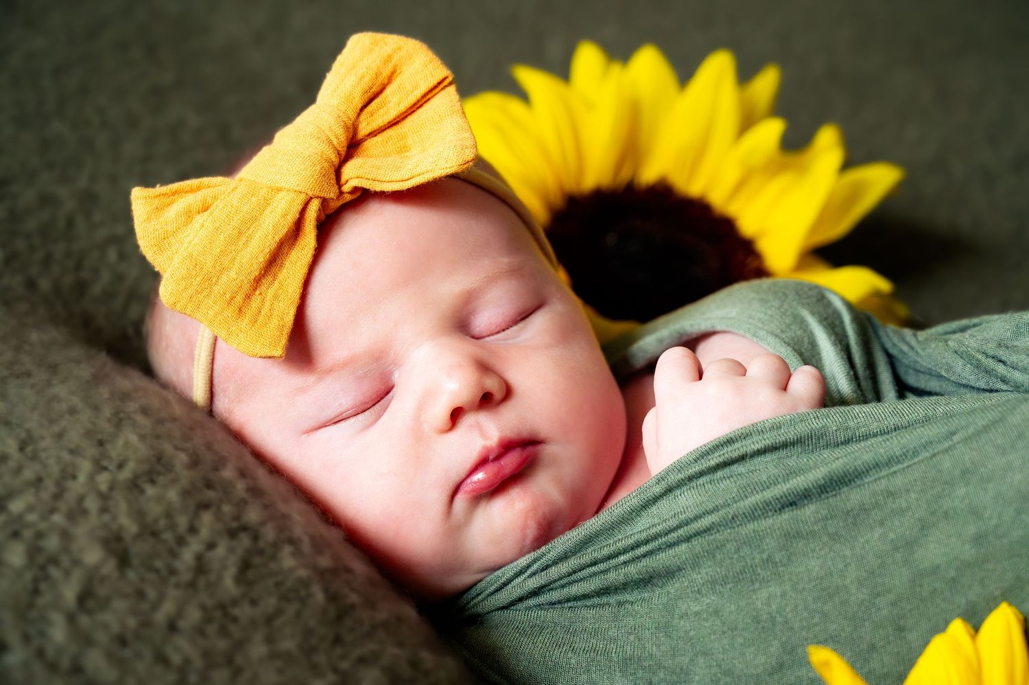 Sleeping baby surrounded by sunflowers and wearing yellow bow.
