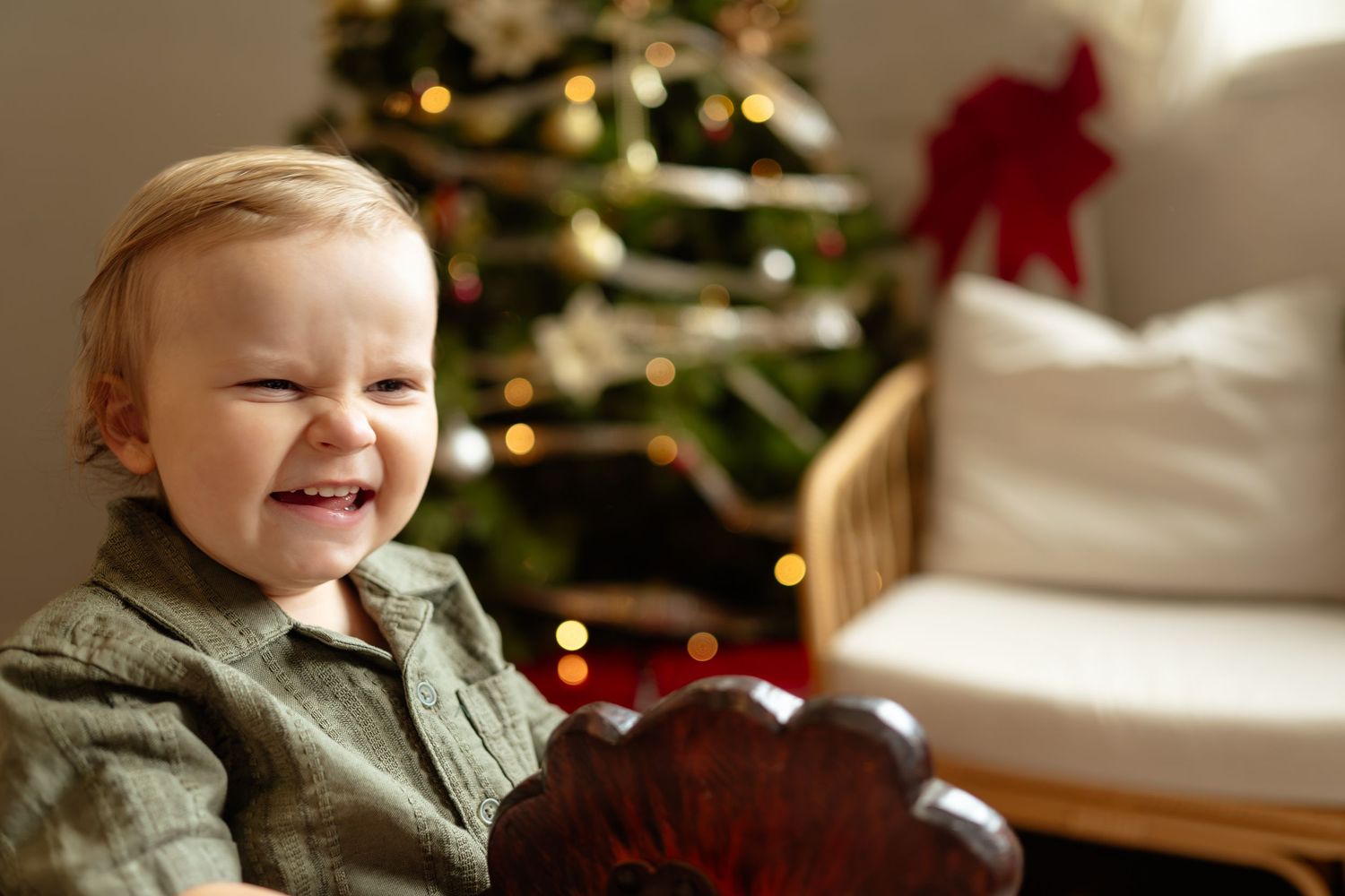 Smiling baby in green outfit sitting near a decorated Christmas tree with bokeh lights.